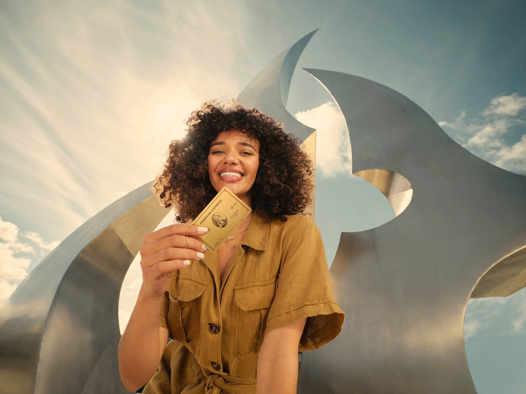 Eine lächelnde junge Frau hält eine goldene Kreditkarte von American Express in der Hand, im Hintergrund eine moderne Skulptur im Freien bei Sonnenschein. Eine lächelnde junge Frau hält eine goldene Kreditkarte von American Express in der Hand, im Hintergrund eine moderne Skulptur im Freien bei Sonnenschein.