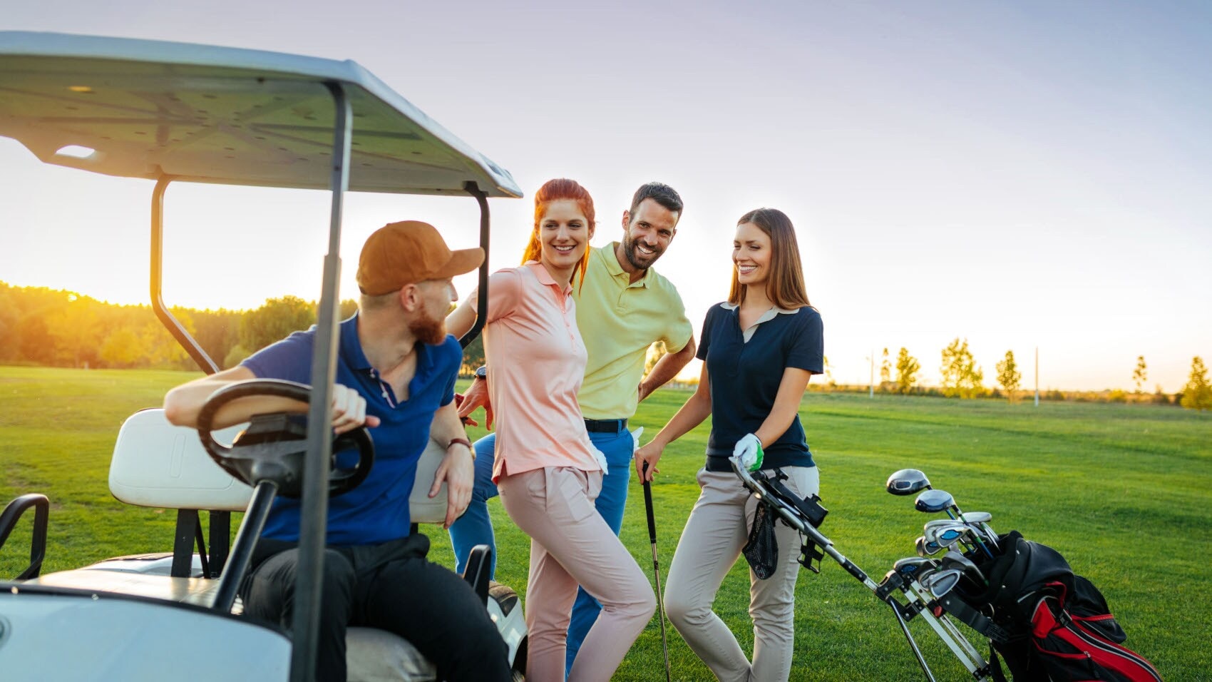 Vier mittelalte Personen in verschiedenfarbigen Polohemden mit Golfequipment und Golfcart stehen beieinander auf einem Golfplatz bei Sonnenuntergang. Vier mittelalte Personen in verschiedenfarbigen Polohemden mit Golfequipment und Golfcart stehen beieinander auf einem Golfplatz bei Sonnenuntergang.