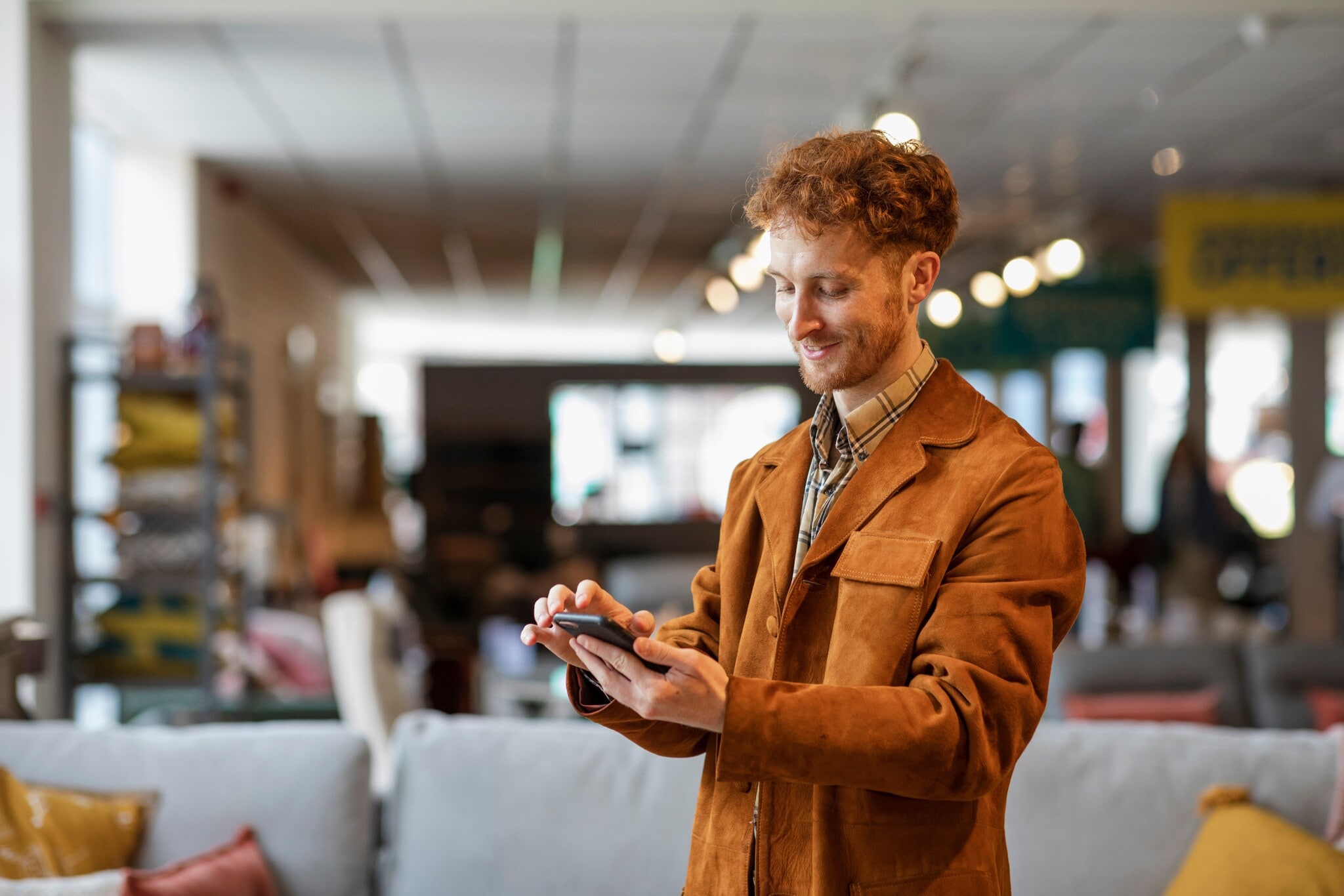 Ein junger Mann steht in einem Möbelgeschäft und schaut lächelnd auf das Smartphone in seiner Hand. Ein junger Mann steht in einem Möbelgeschäft und schaut lächelnd auf das Smartphone in seiner Hand.