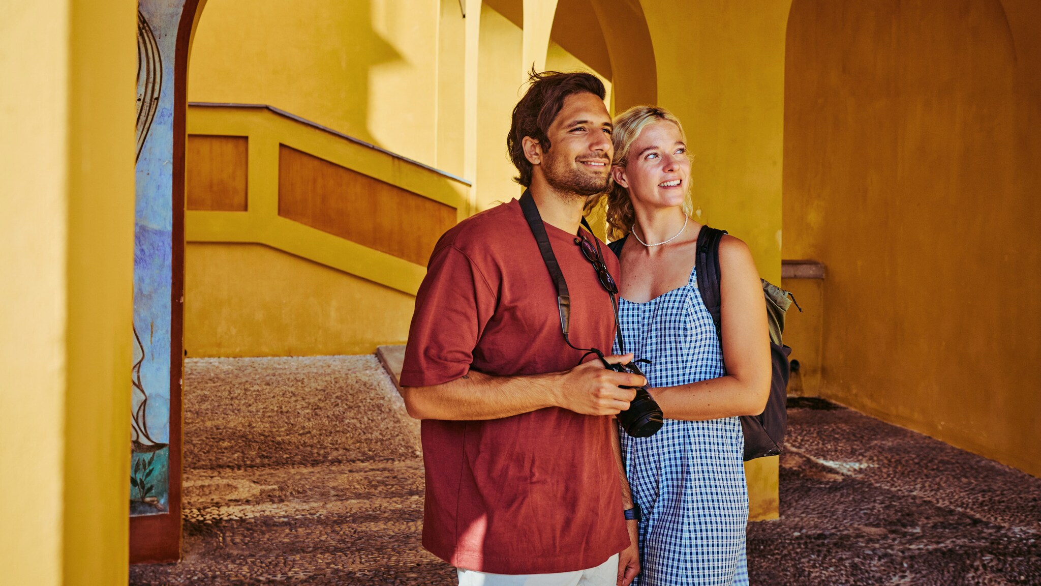 Ein Mann und eine Frau in Sommerkleidung mit Rucksack und Kamera vor einer gelben Hausfassade, die in dieselbe Richtung blicken.