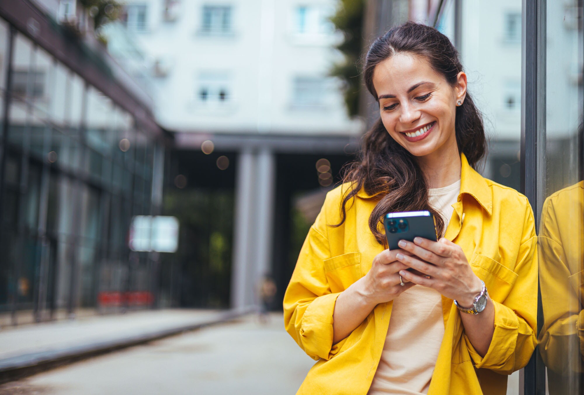 Eine Frau im gelben Hemd lehnt an einer Außenfassade und schaut lächelnd auf das Smartphone in ihren Händen. Eine Frau im gelben Hemd lehnt an einer Außenfassade und schaut lächelnd auf das Smartphone in ihren Händen.