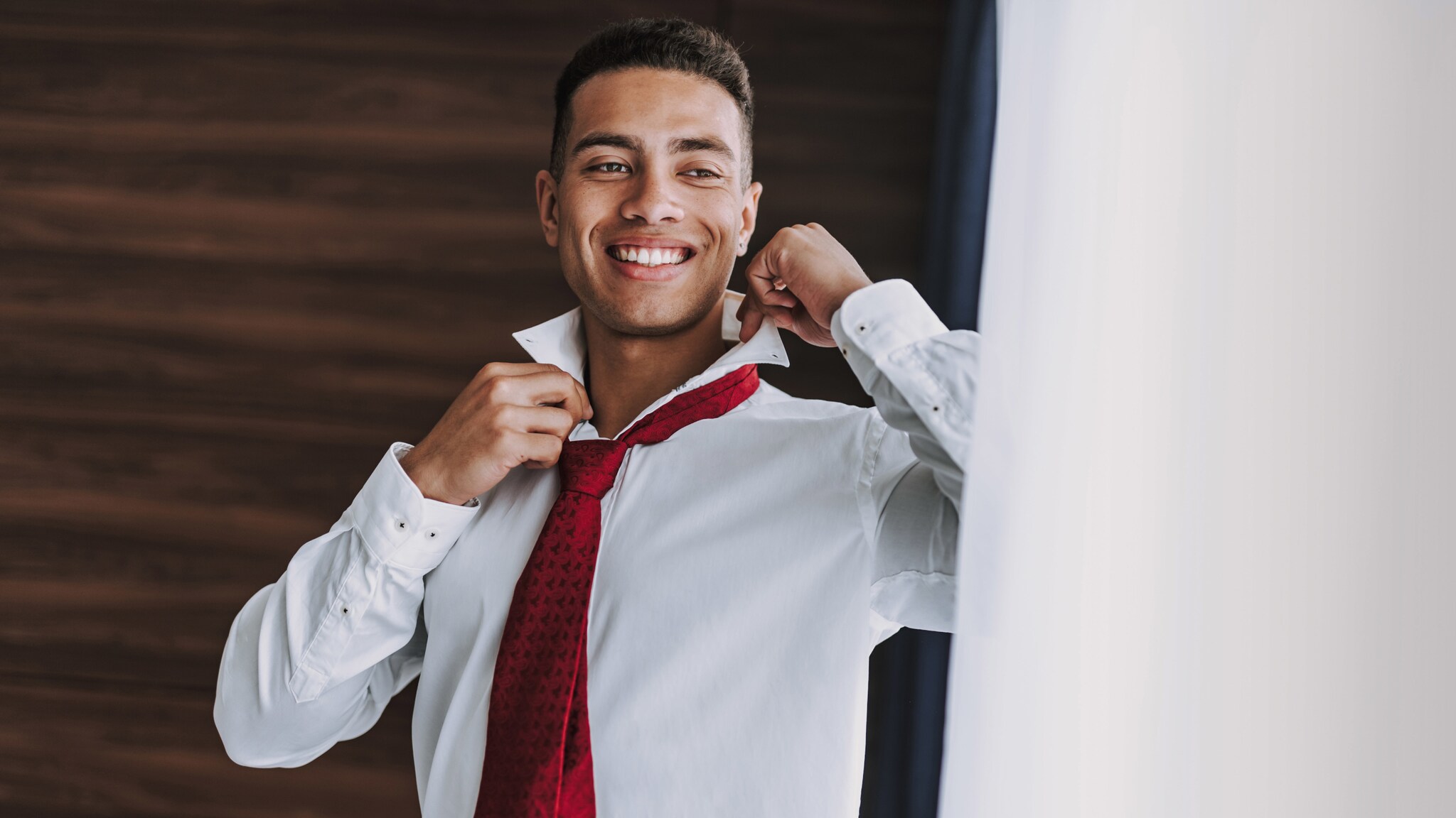 Ein junger Mann im weißen Hemd mit roter Krawatte richtet sich lächelnd den Kragen vor einem Spiegel. Ein junger Mann im weißen Hemd mit roter Krawatte richtet sich lächelnd den Kragen vor einem Spiegel.