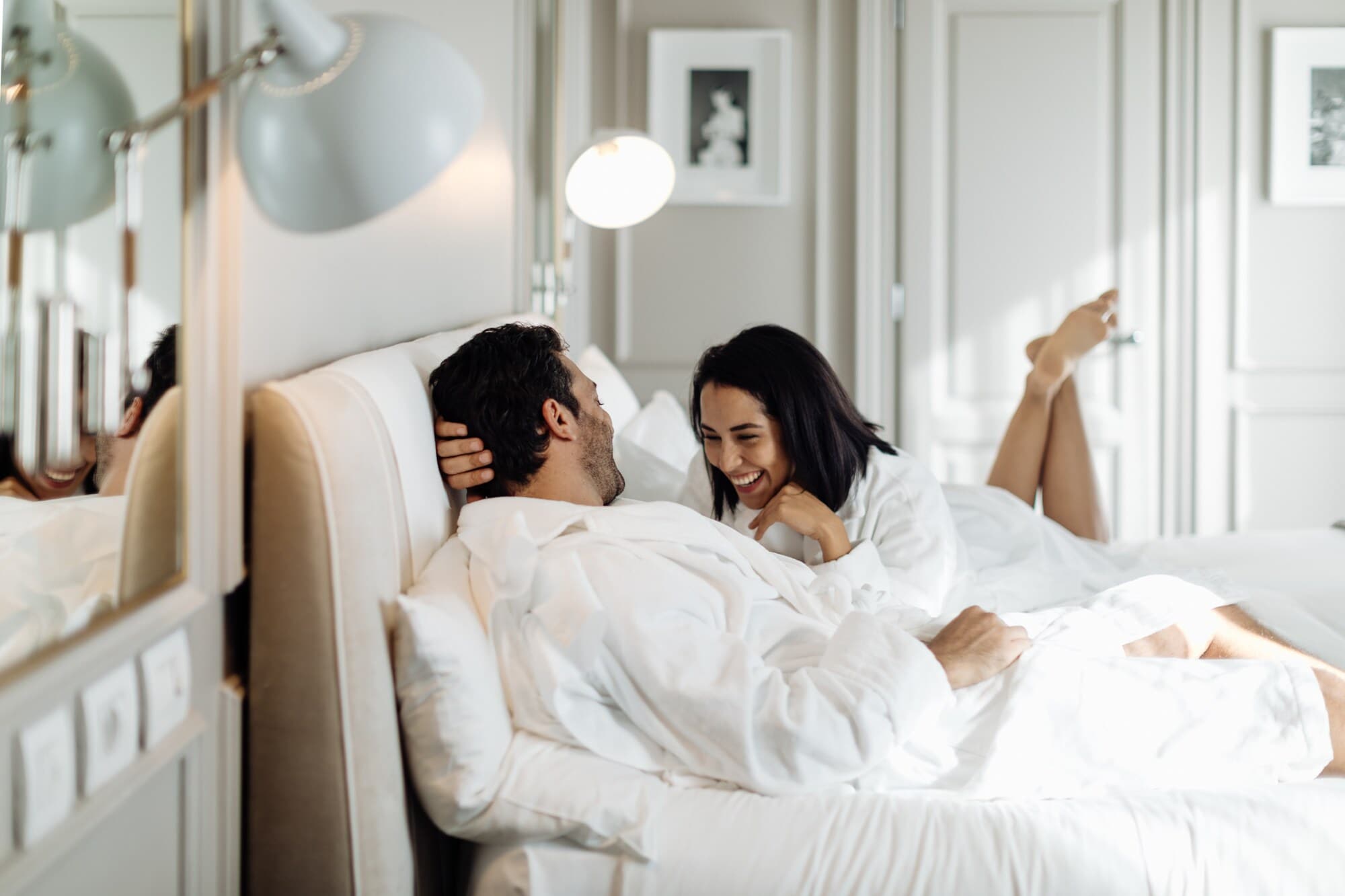 Ein Paar in weißen Bademänteln liegt lachend in einem weißen Bett in einem edlen, hellen Zimmer. Ein Paar in weißen Bademänteln liegt lachend in einem weißen Bett in einem edlen, hellen Zimmer.
