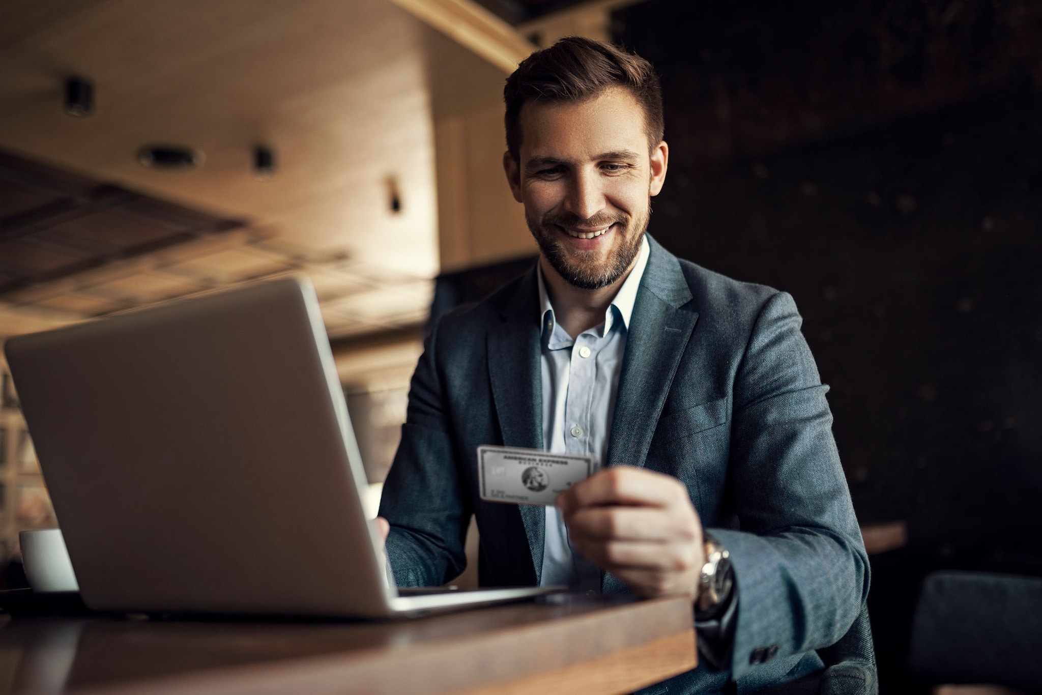 American Express Business Platinum Card: Unternehmen online managen Ein lächelnder Geschäftsmann im Anzug sitzt an einem Tisch vor einem Laptop und blickt auf die Platinum Card von American Express in seiner Hand.