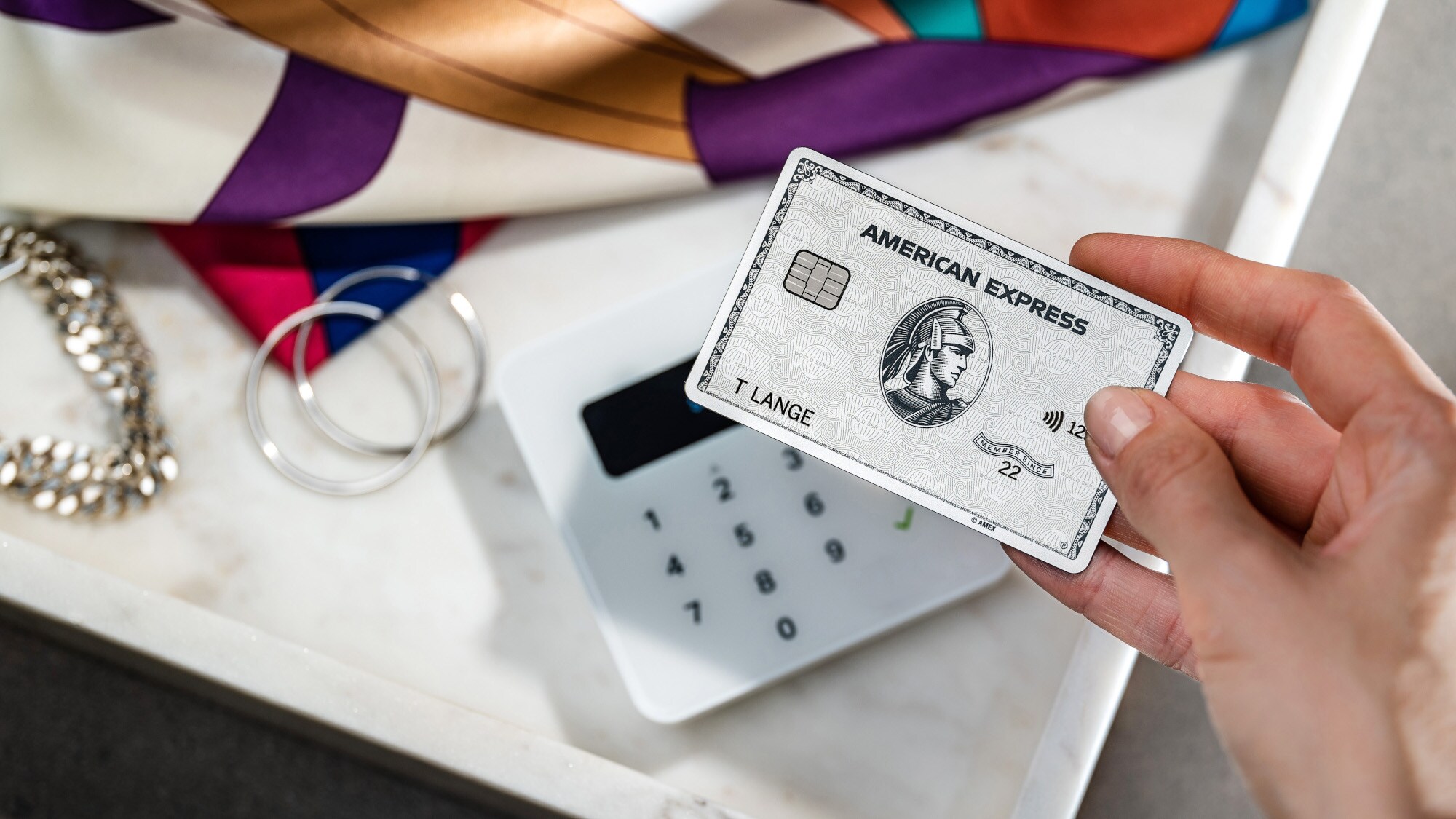 Nahaufnahme einer Hand, die eine silberne American Express Kreditkarte an ein Kartenlesegerät neben Modeaccessoires hält. Nahaufnahme einer Hand, die eine silberne American Express Kreditkarte an ein Kartenlesegerät neben Modeaccessoires hält.