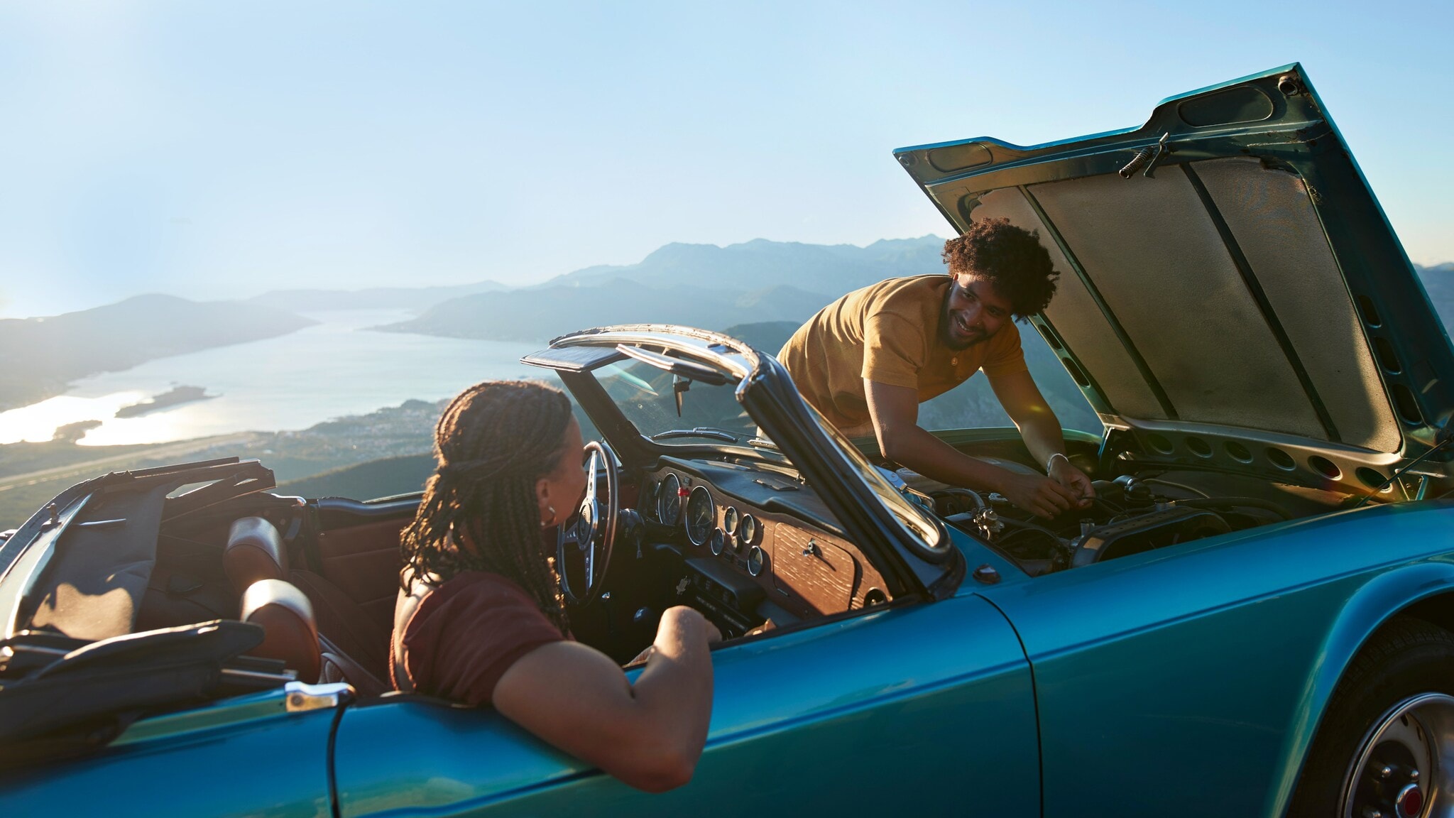 Ein Paar in einem liegengebliebenen Cabriolet an einer Meeresküste bei Sonnenschein, er kontrolliert den Motor, sie sitzt auf dem Beifahrersitz. Ein Paar in einem liegengebliebenen Cabriolet an einer Meeresküste bei Sonnenschein, er kontrolliert den Motor, sie sitzt auf dem Beifahrersitz.