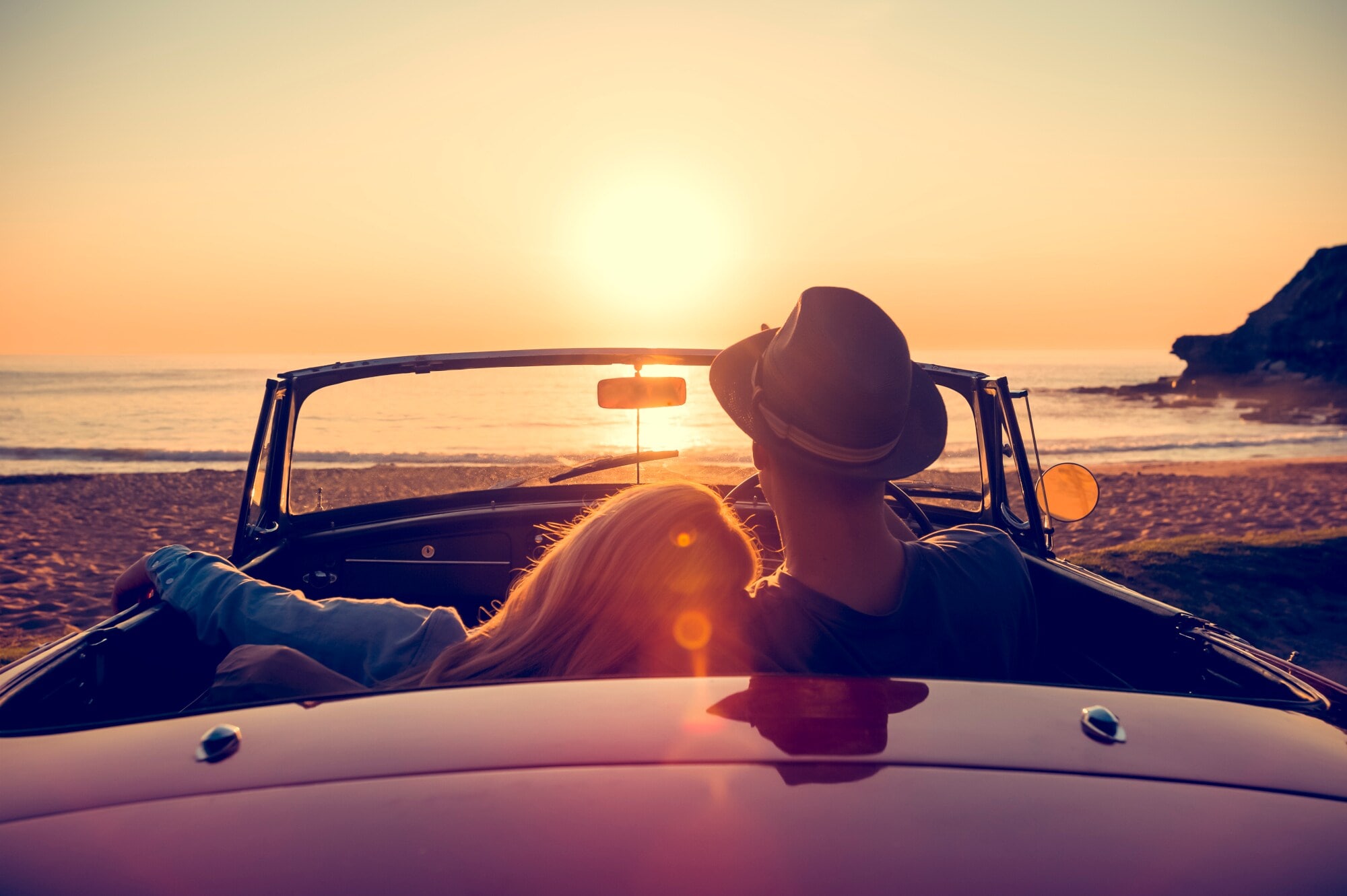 Rückansicht eines Paares in einem Cabriolet an einem Sandstrand bei Sonnenuntergang, sie hat den Kopf auf seine Schulter gelehnt. Rückansicht eines Paares in einem Cabriolet an einem Sandstrand bei Sonnenuntergang, sie hat den Kopf auf seine Schulter gelehnt.