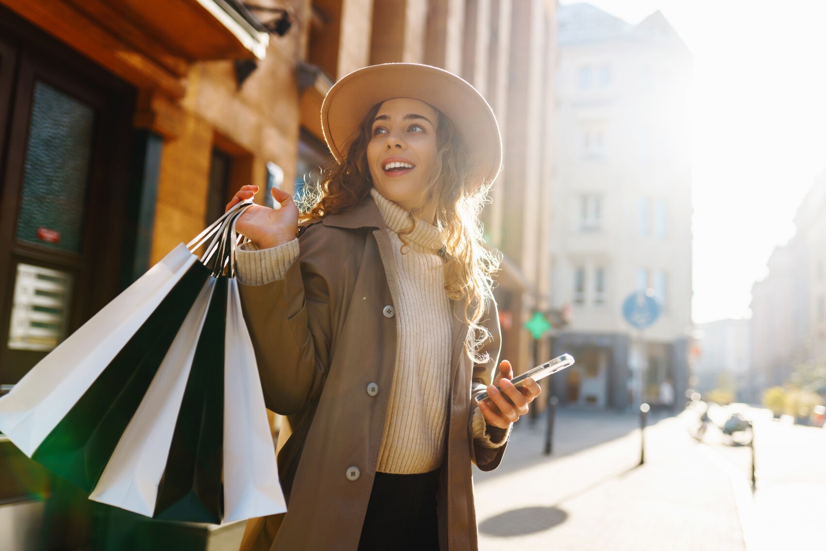 Eine junge Frau mit Hut steht in einer Innenstadt und hält ein Smartphone und mehrere Einkaufstüten in den Händen.