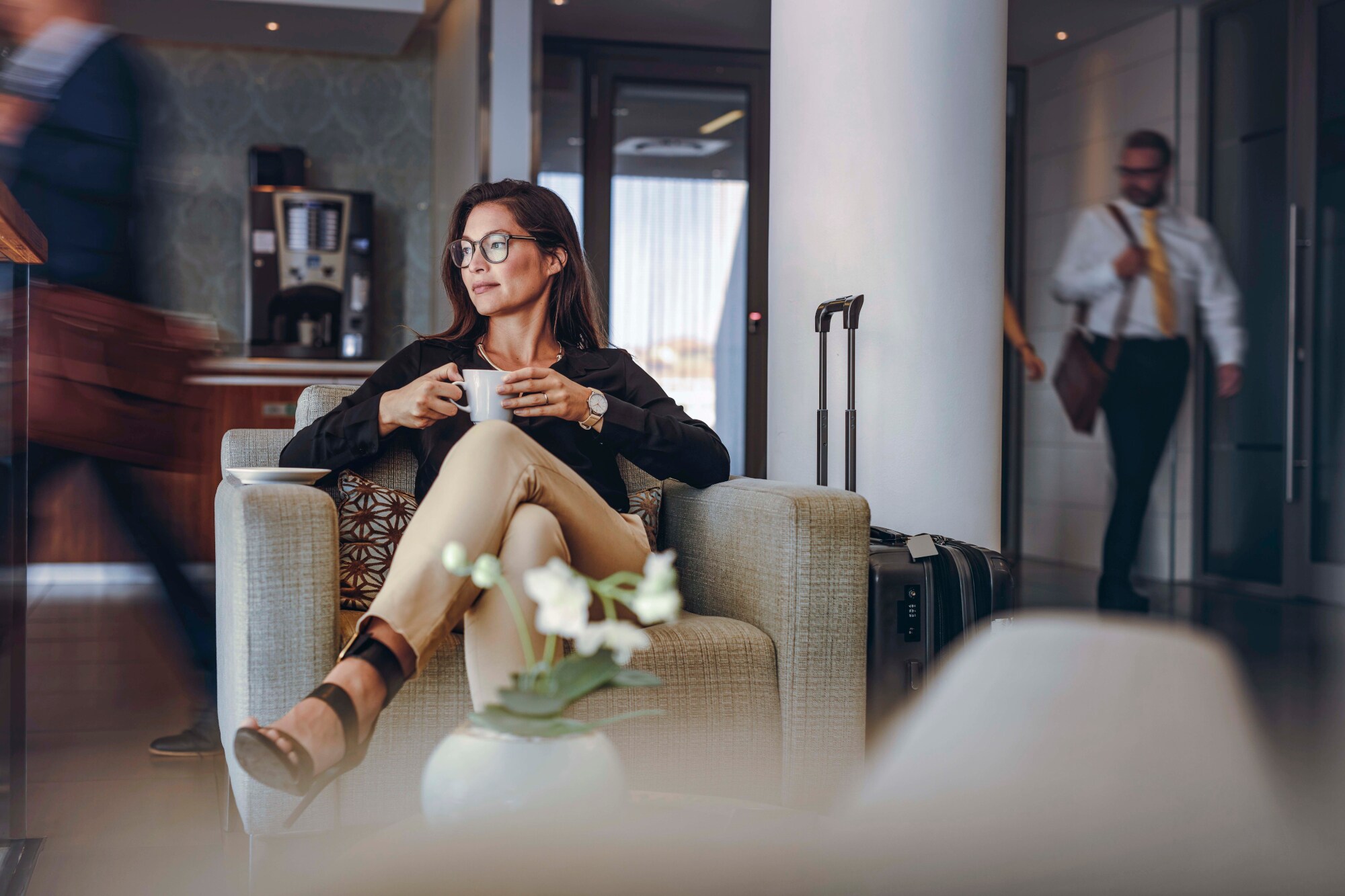 Eine Frau sitzt mit einer Kaffeetasse in einem Sessel in einer belebten Lounge, neben ihr ein Reisekoffer.