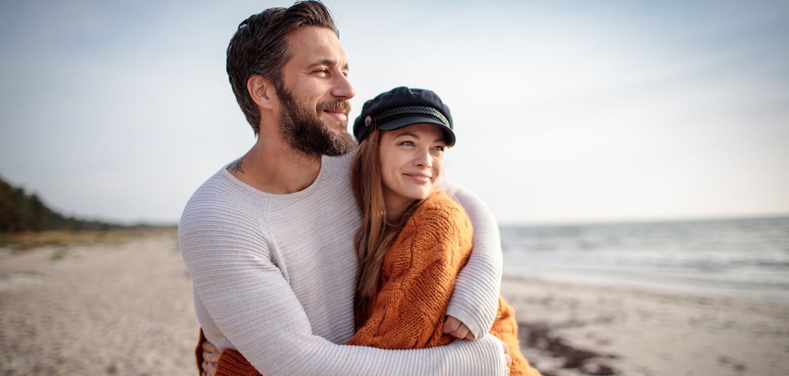 Ein sich umarmendes Paar in Pullovern steht glücklich lächelnd an einem Sandstrand und blickt aufs Meer. Ein sich umarmendes Paar in Pullovern steht glücklich lächelnd an einem Sandstrand und blickt aufs Meer.