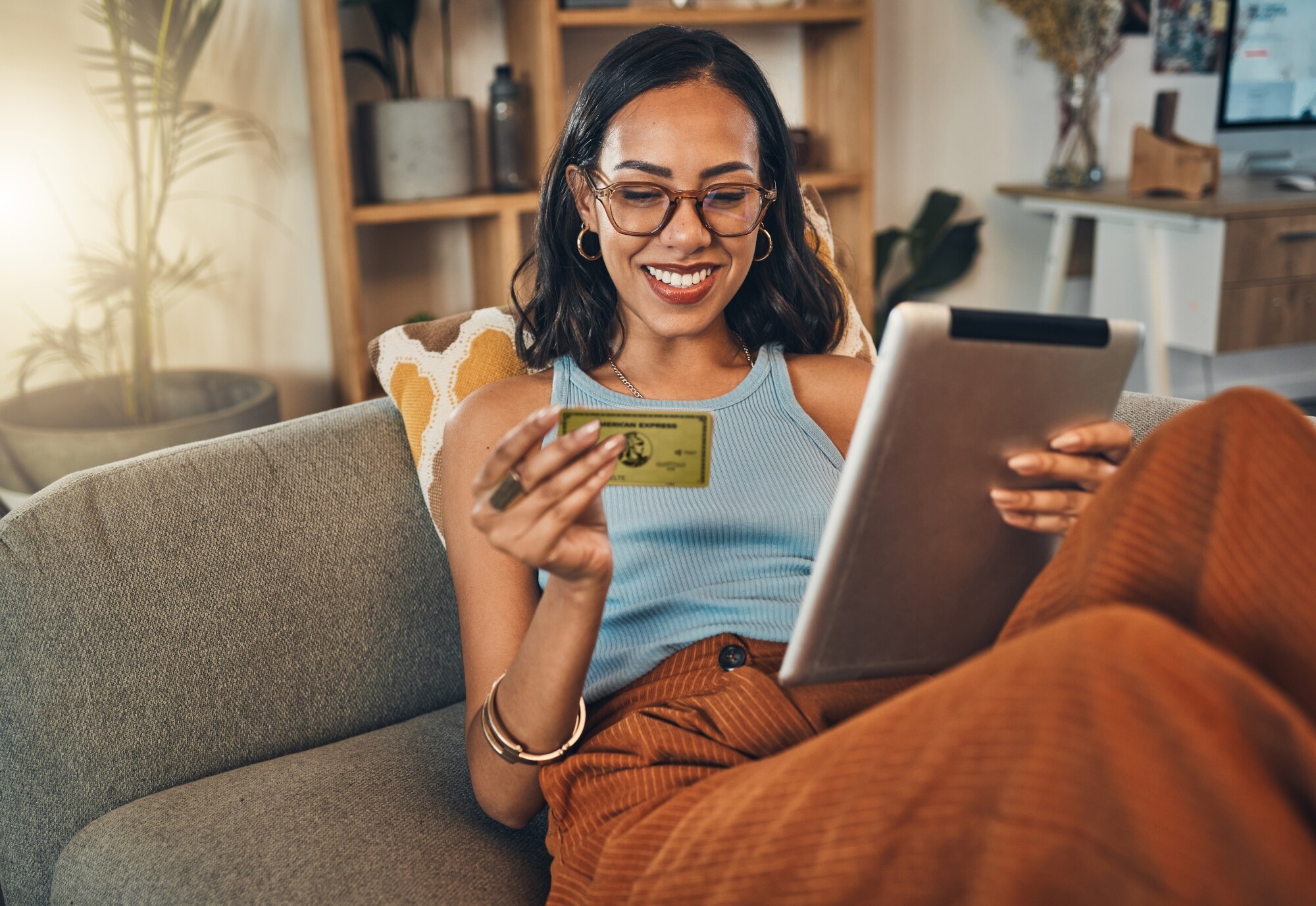 Eine junge Frau sitzt lächelnd auf einem Sofa, in ihren Händen hält sie ein Tablet und eine goldene Kreditkarte von American Express.