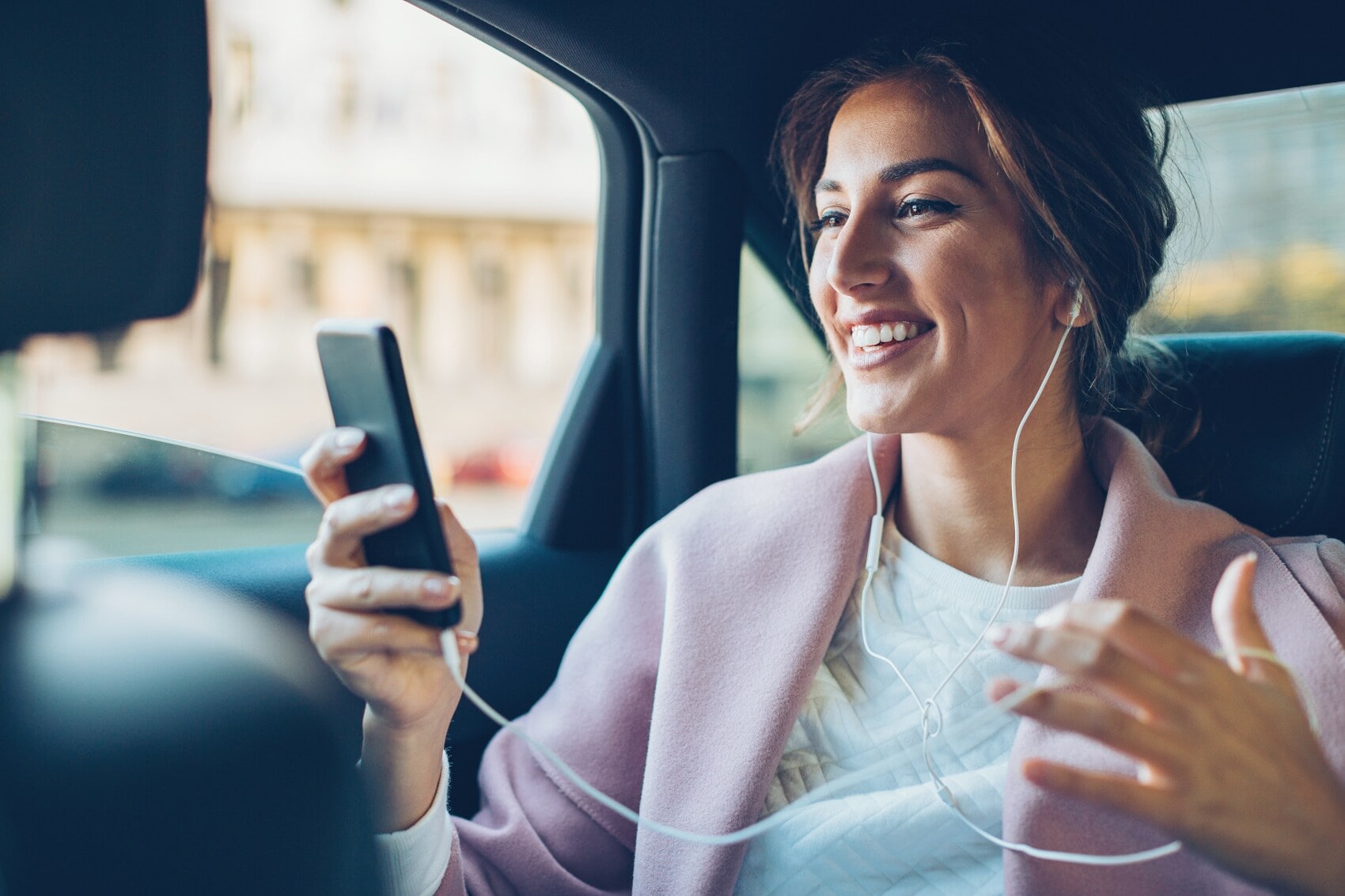 Eine junge Frau mit Kopfhörern auf dem Rücksitz eines Autos schaut lachend auf das Display ihres Smartphones. Eine junge Frau mit Kopfhörern auf dem Rücksitz eines Autos schaut lachend auf das Display ihres Smartphones.