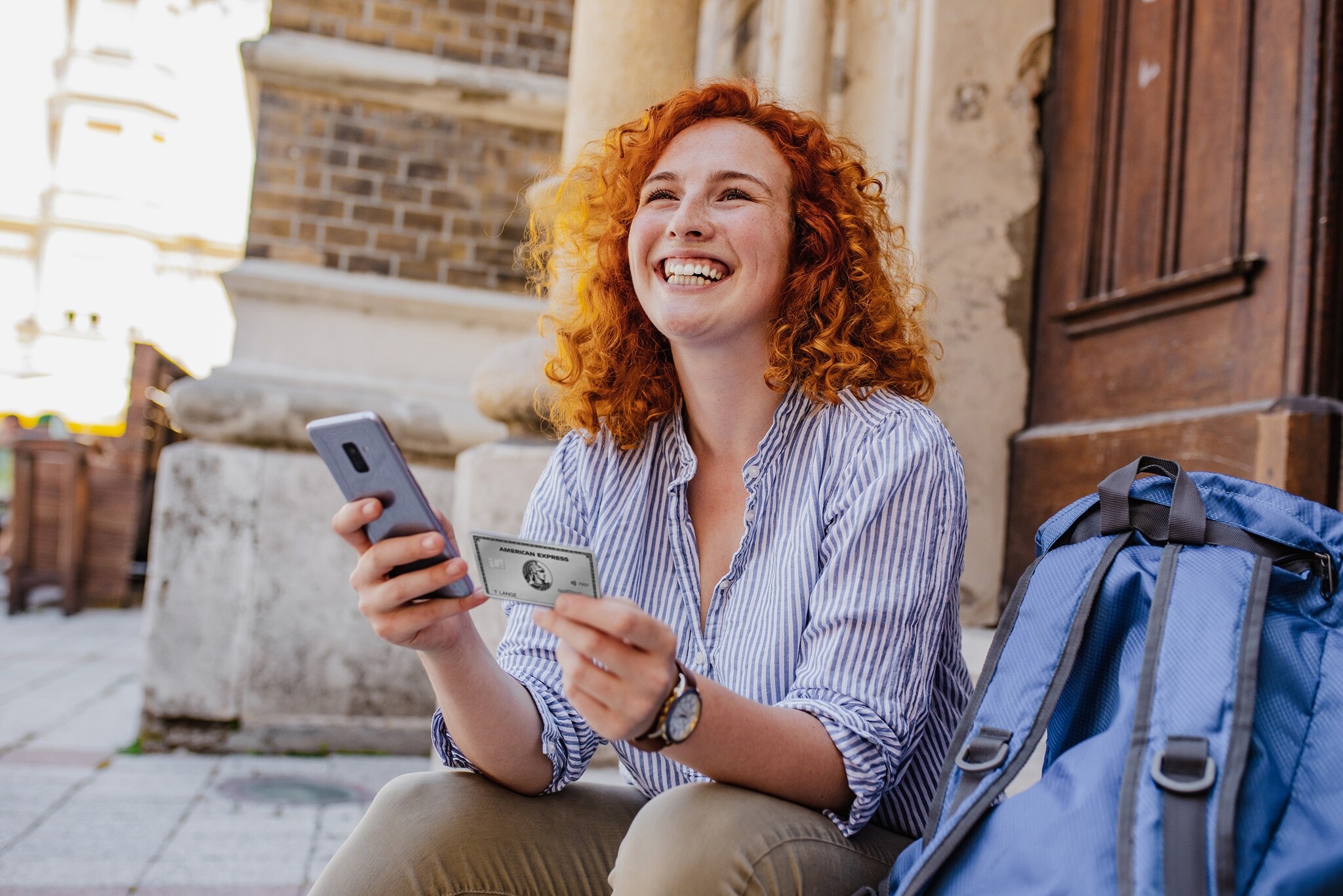 Eine junge Frau mit Smartphone und Kreditkarte in den Händen sitzt gut gelaunt auf einer Treppenstufe am Eingang eines Gebäudes, neben ihr ein Rucksack. Eine junge Frau mit Smartphone und Kreditkarte in den Händen sitzt gut gelaunt auf einer Treppenstufe am Eingang eines Gebäudes, neben ihr ein Rucksack.