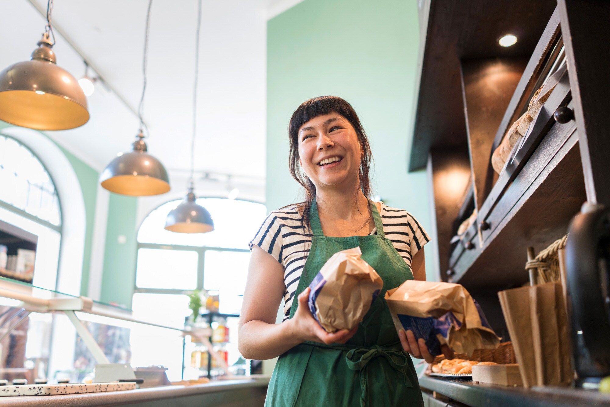 Eine Verkäuferin mit Backwaren in den Händen steht lachend hinter der Theke einer Bäckerei. Eine Verkäuferin mit Backwaren in den Händen steht lachend hinter der Theke einer Bäckerei.