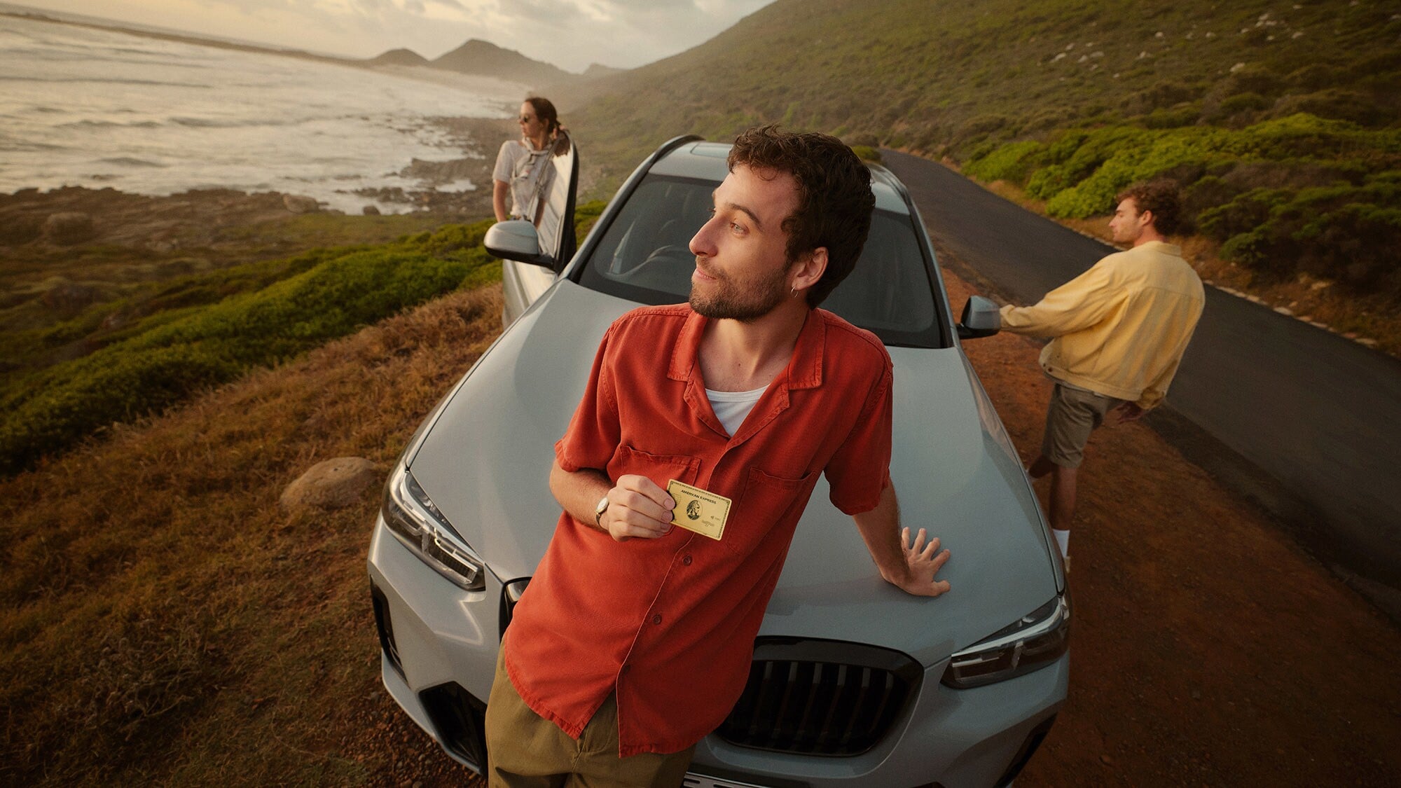 Ein junger Mann lehnt an der Kühlerhaube eines Autos in einer Küstenlandschaft und blickt aufs Meer, in einer Hand hält er eine goldene Kreditkarte von American Express, dahinter zwei weitere Personen am Auto. Ein junger Mann lehnt an der Kühlerhaube eines Autos in einer Küstenlandschaft und blickt aufs Meer, in einer Hand hält er eine goldene Kreditkarte von American Express, dahinter zwei weitere Personen am Auto.