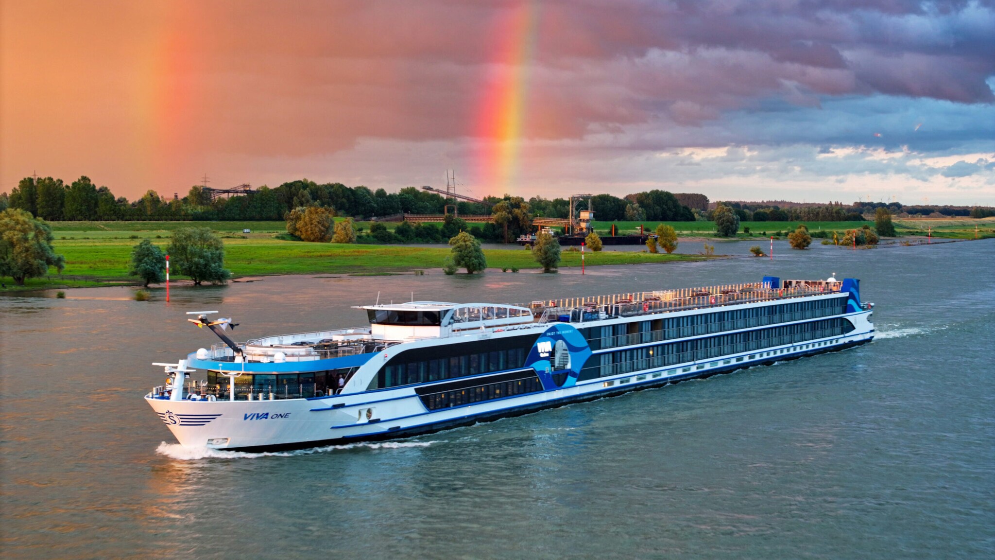 Flusskreuzfahrtschiff Viva One fährt vor grünem Flussufer mit Regenbogen am bewölkten Himmel. Flusskreuzfahrtschiff Viva One fährt vor grünem Flussufer mit Regenbogen am bewölkten Himmel.