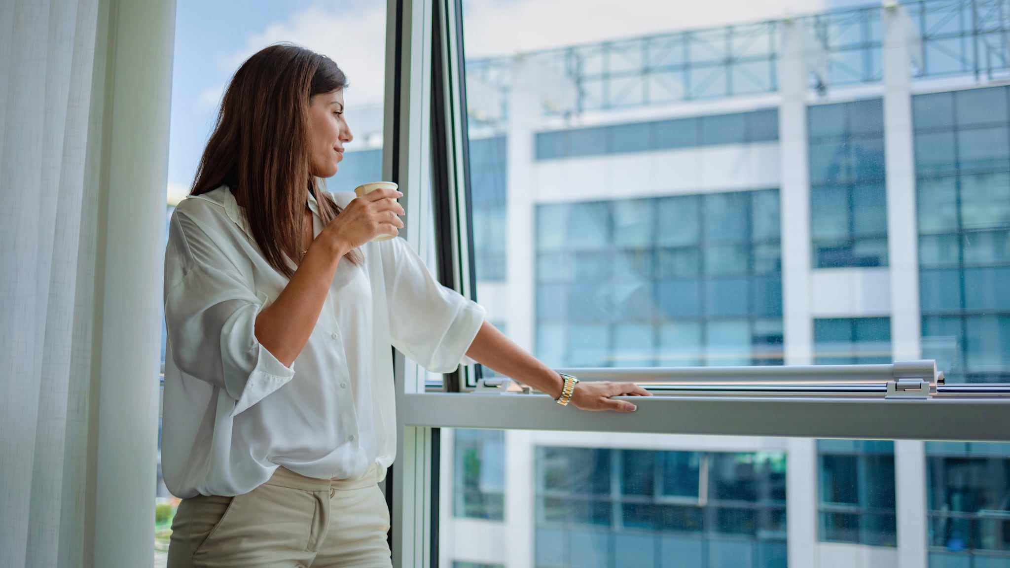 Eine Frau in weißer Bluse steht mit einem Kaffeebecher in der Hand an einem halbgeöffneten Fenster und blickt hinaus auf ein gläsernes Gebäude.