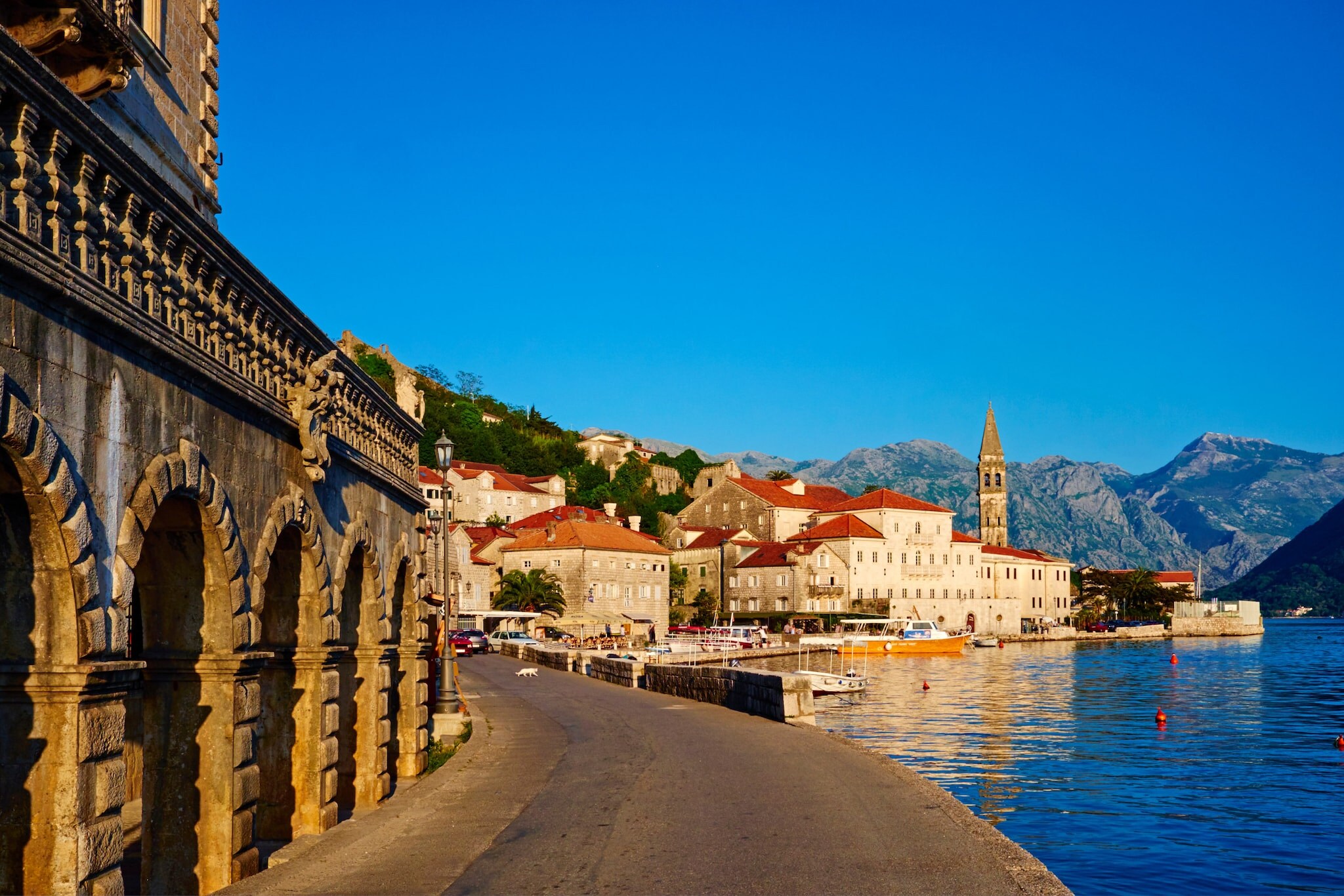 Häuser der Stadt Perast am Wasser, im Hintergrund Berge und blauer Himmel.
