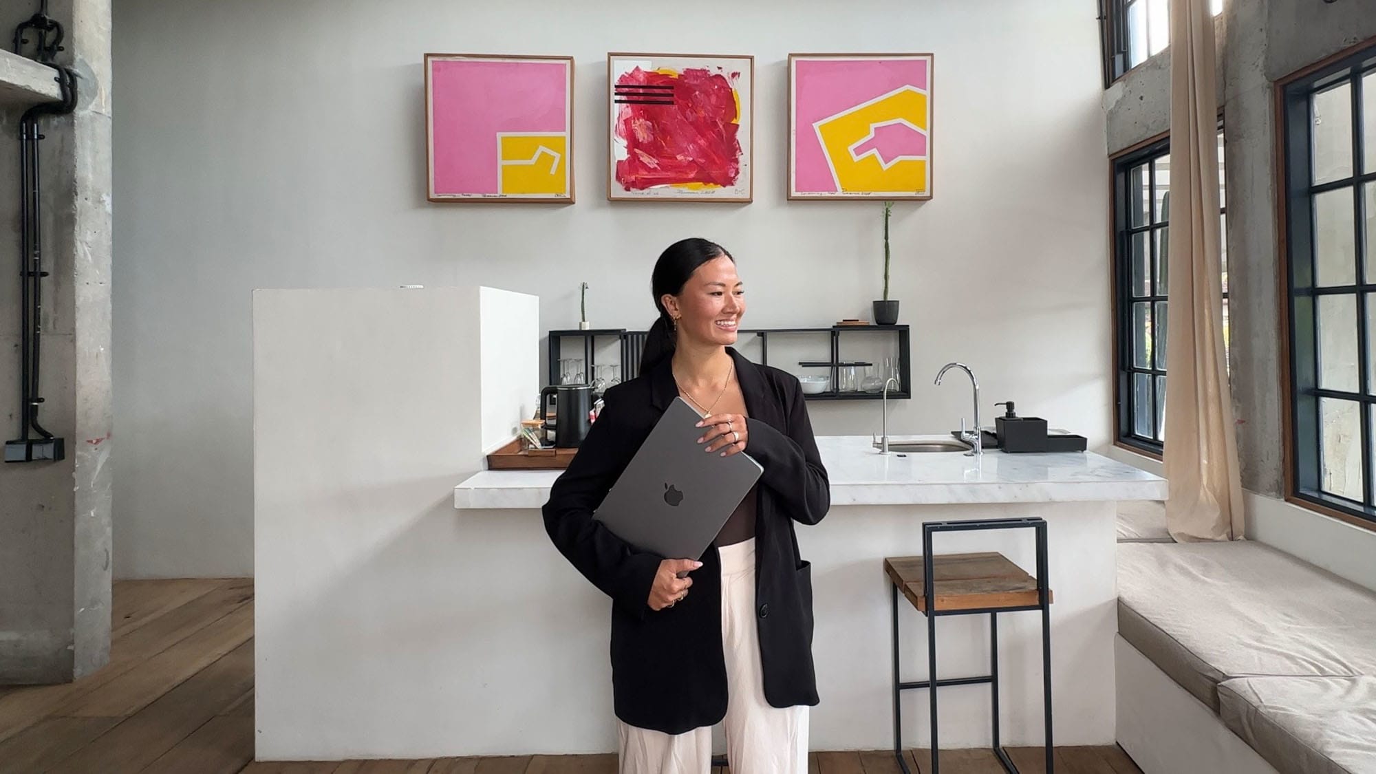 Eine lächelnde Frau mit Laptop unter dem Arm steht vor einer modernen Küchenzeile mit abstrakten Gemälden in einem Loft. Eine lächelnde Frau mit Laptop unter dem Arm steht vor einer modernen Küchenzeile mit abstrakten Gemälden in einem Loft.