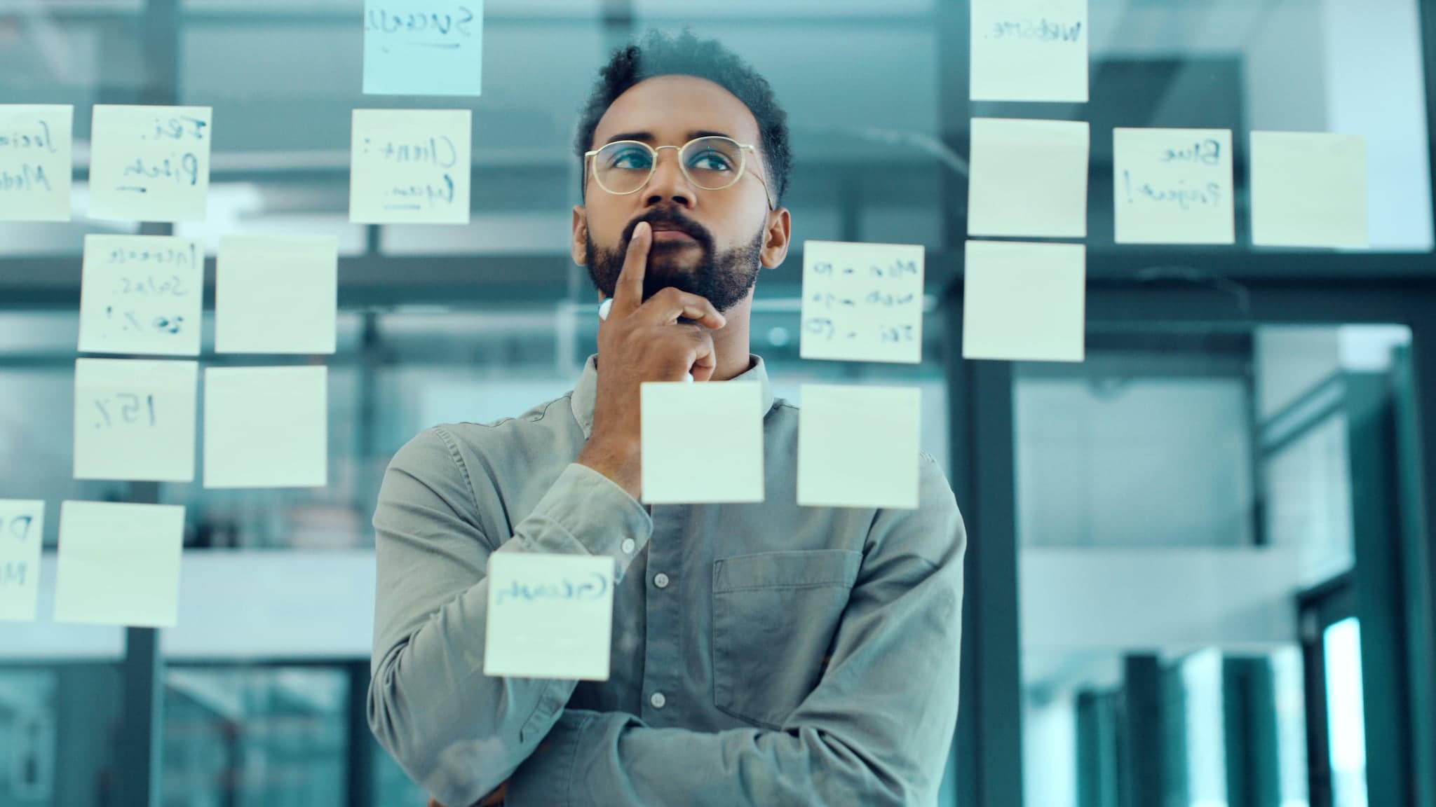 Ein Mann mit einem Finger an der Lippe steht hinter einer Glasscheibe mit beschriebenen Post-its. Ein Mann mit einem Finger an der Lippe steht hinter einer Glasscheibe mit beschriebenen Post-its.