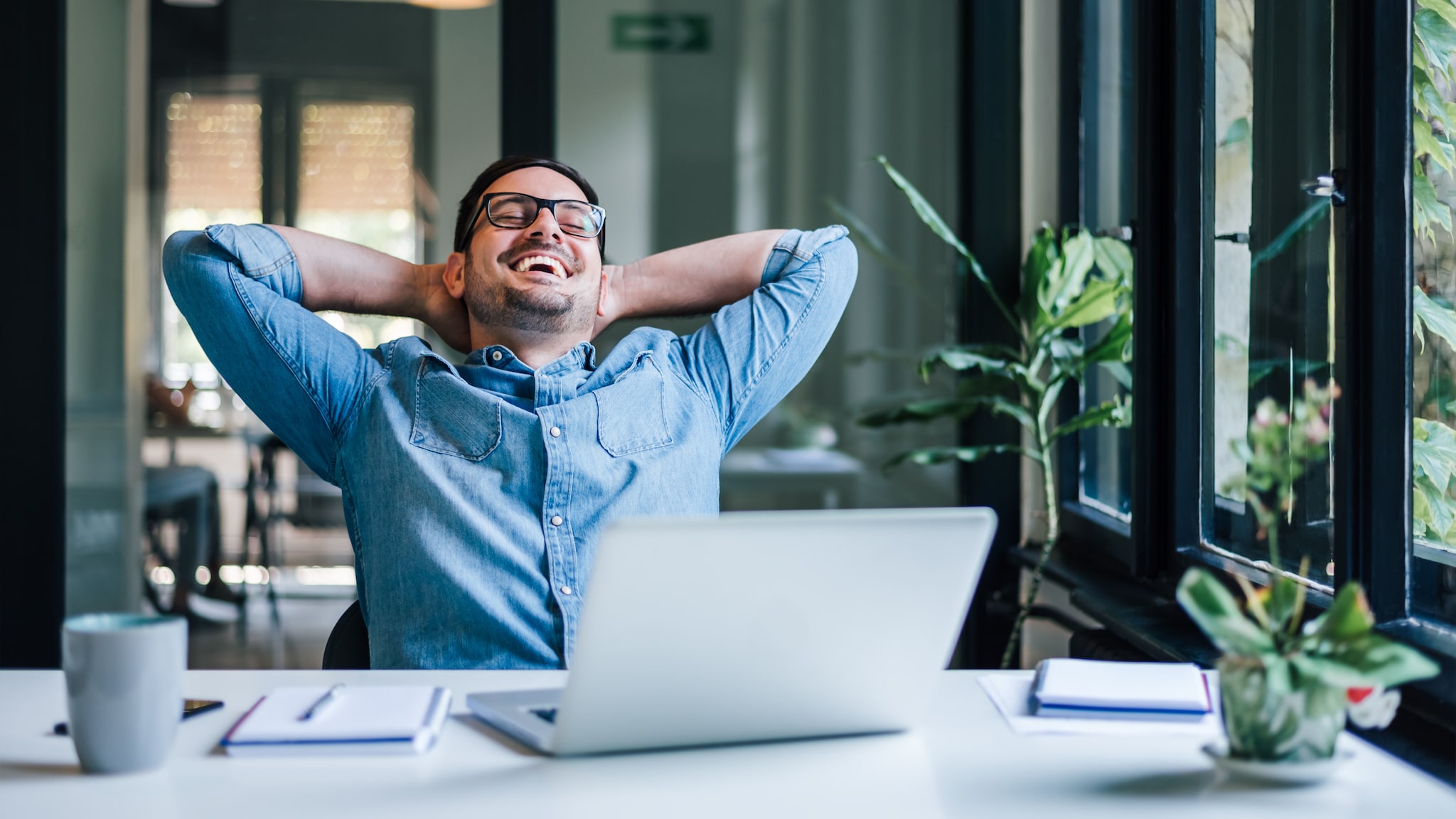 Ein lachender Mann lehnt entspannt zurück mit verschränkten Armen im Nacken auf einem Stuhl an einem Schreibtisch.