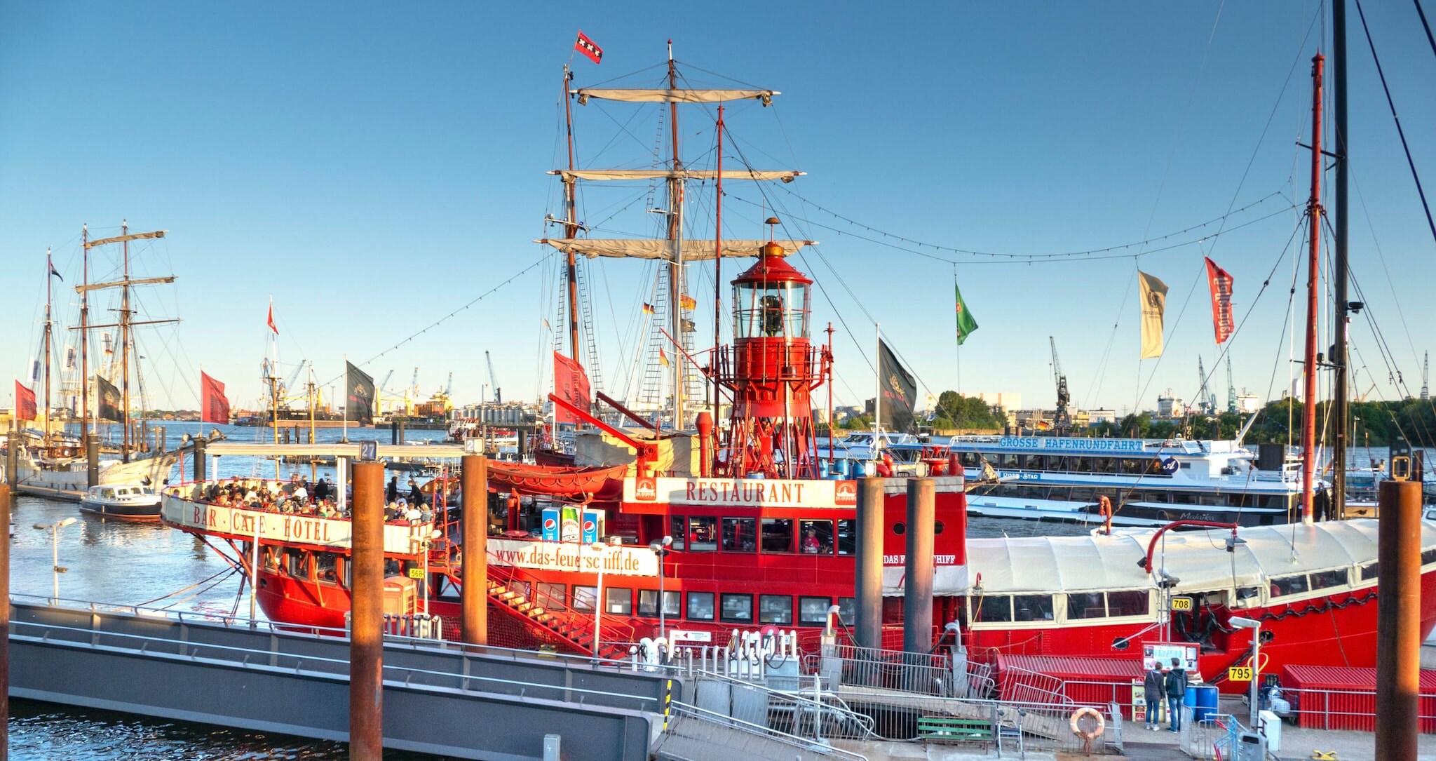 Personen an Deck eines roten, historischen Feuerschiffes mit Gastronomiebetrieb im Hafen.