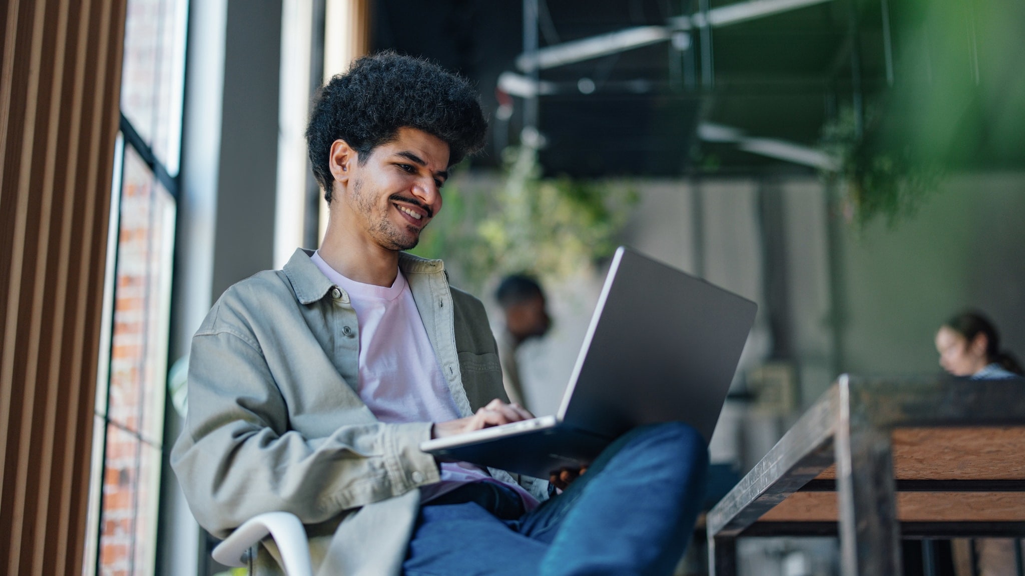 Ein junger Mann mit aufgeklapptem Laptop auf seinem Schoß in einem hellen Loft mit weiteren Personen. Ein junger Mann mit aufgeklapptem Laptop auf seinem Schoß in einem hellen Loft mit weiteren Personen.