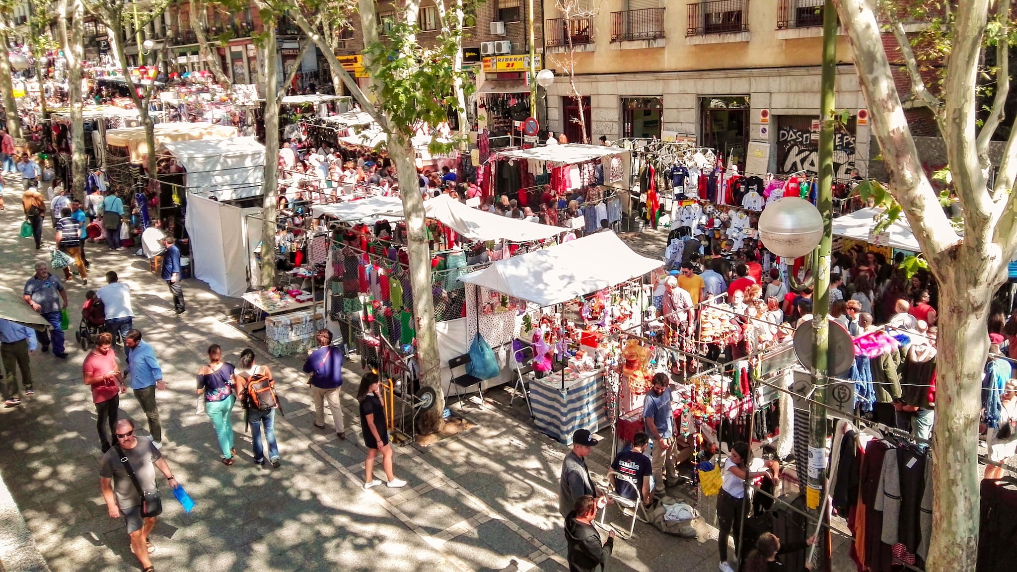Zahlreiche Personen zwischen Flohmarktständen in einer Straße mit Platanen.