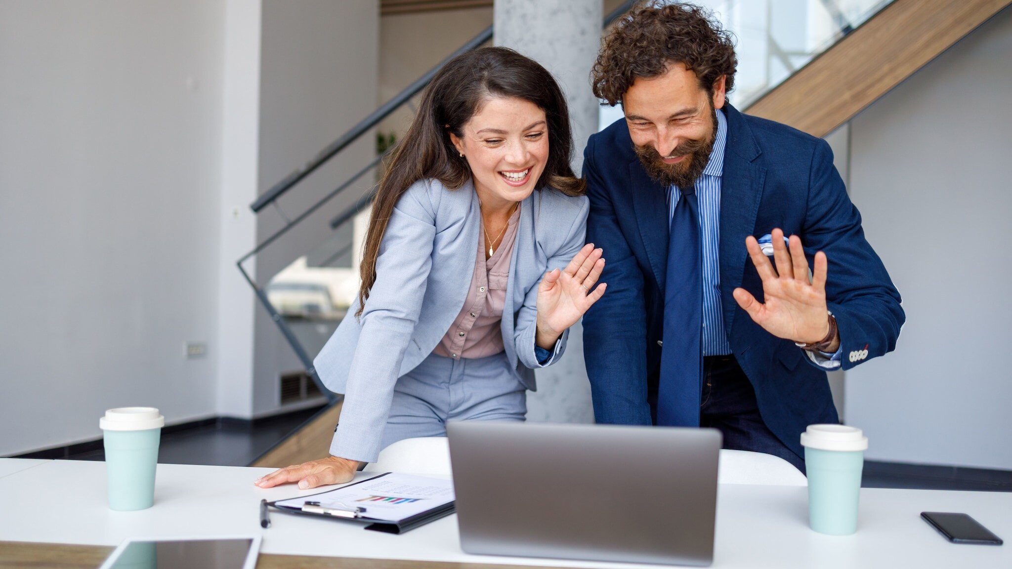 Eine Frau und ein Mann in Business-Kleidung winken lachend gemeinsam in einen Laptop, der vor ihnen auf einem Tisch steht.