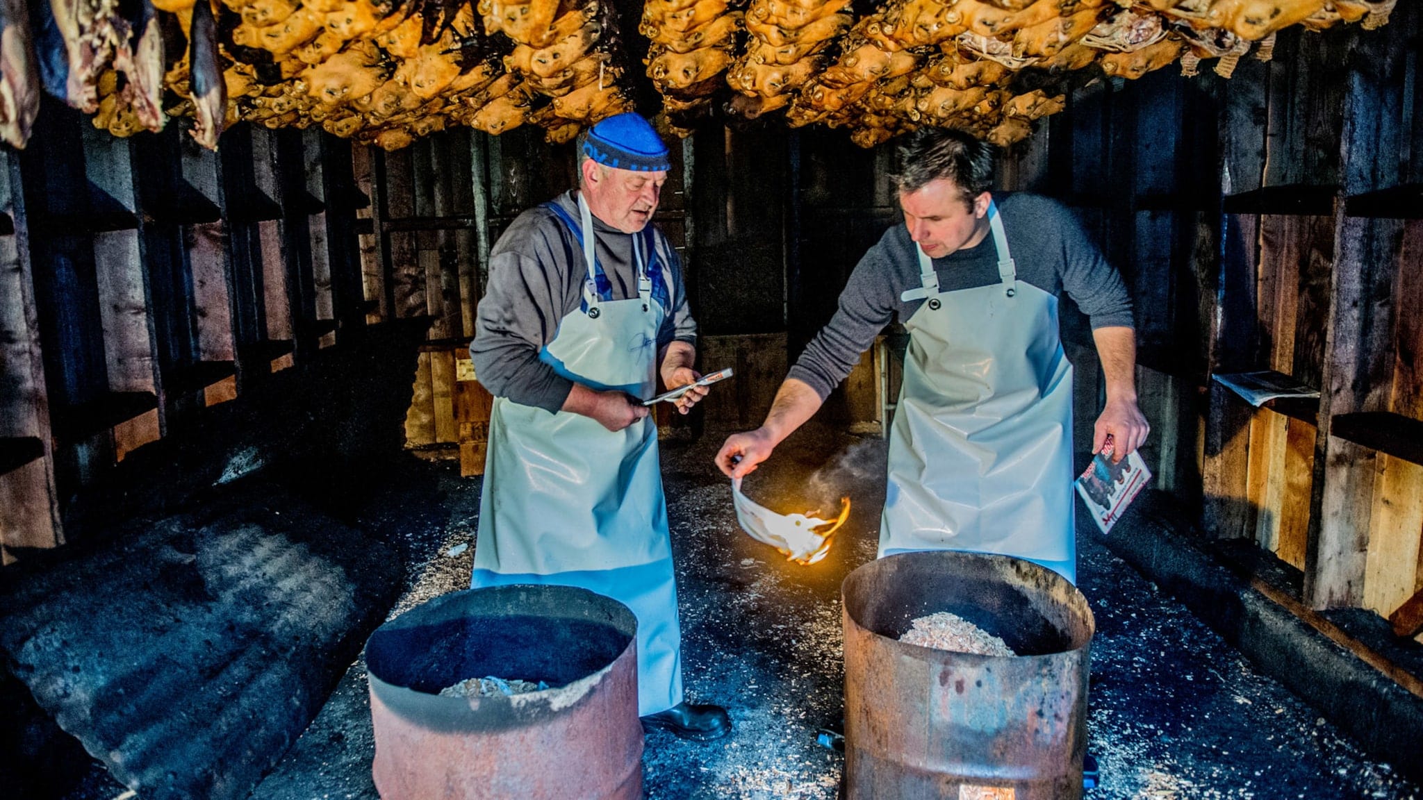 Zwei Männer in Schürzen entzünden ein Feuer an Tonnen in einer Holzhütte mit aufgehängten Schafsköpfen an der Decke. Zwei Männer in Schürzen entzünden ein Feuer an Tonnen in einer Holzhütte mit aufgehängten Schafsköpfen an der Decke.