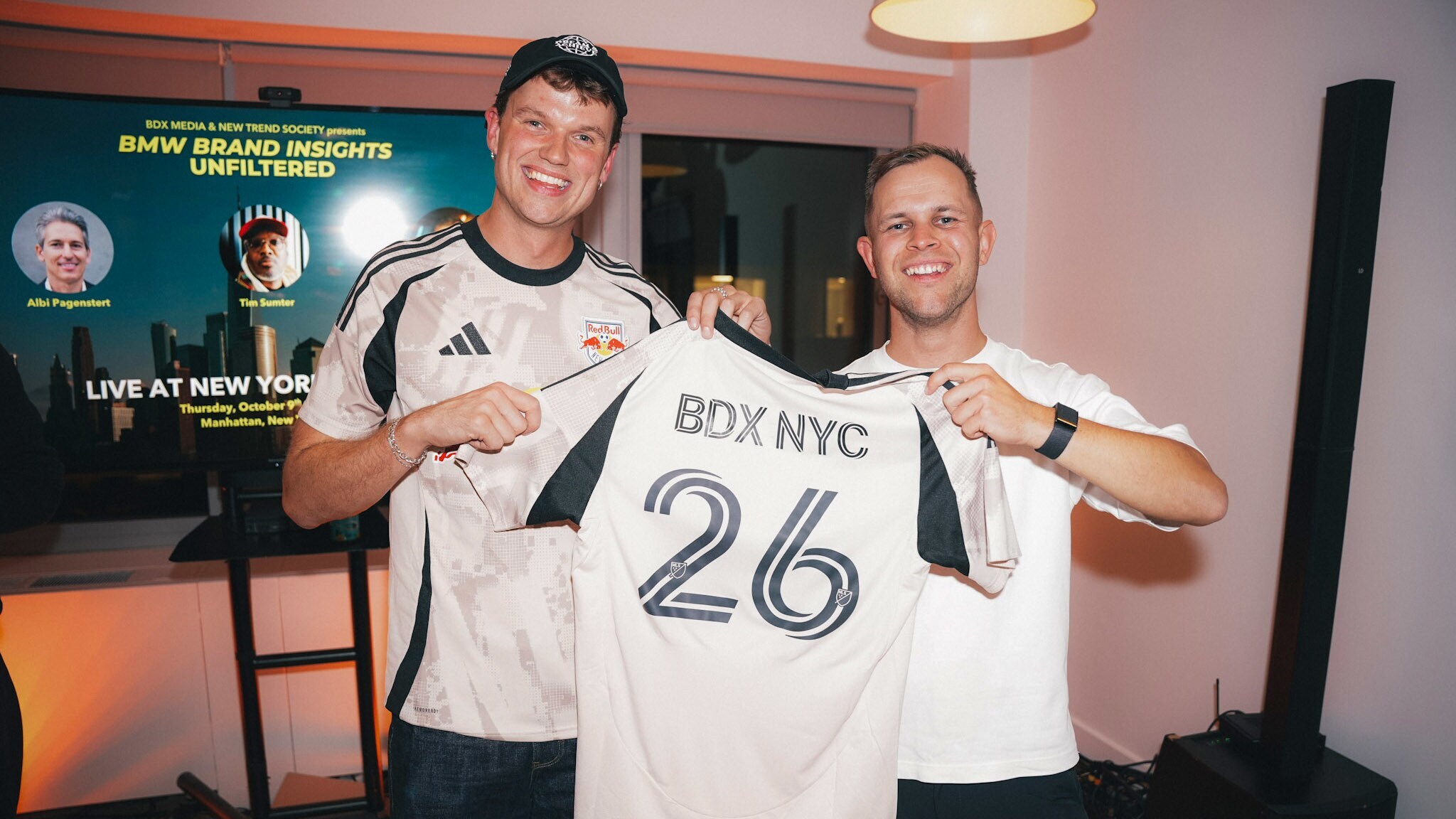 Zwei Männer posieren mit einem Fußballtrikot mit der Aufschrift BDX NYC 26 vor einem Display in einem Raum. Zwei Männer posieren mit einem Fußballtrikot mit der Aufschrift BDX NYC 26 vor einem Display in einem Raum.