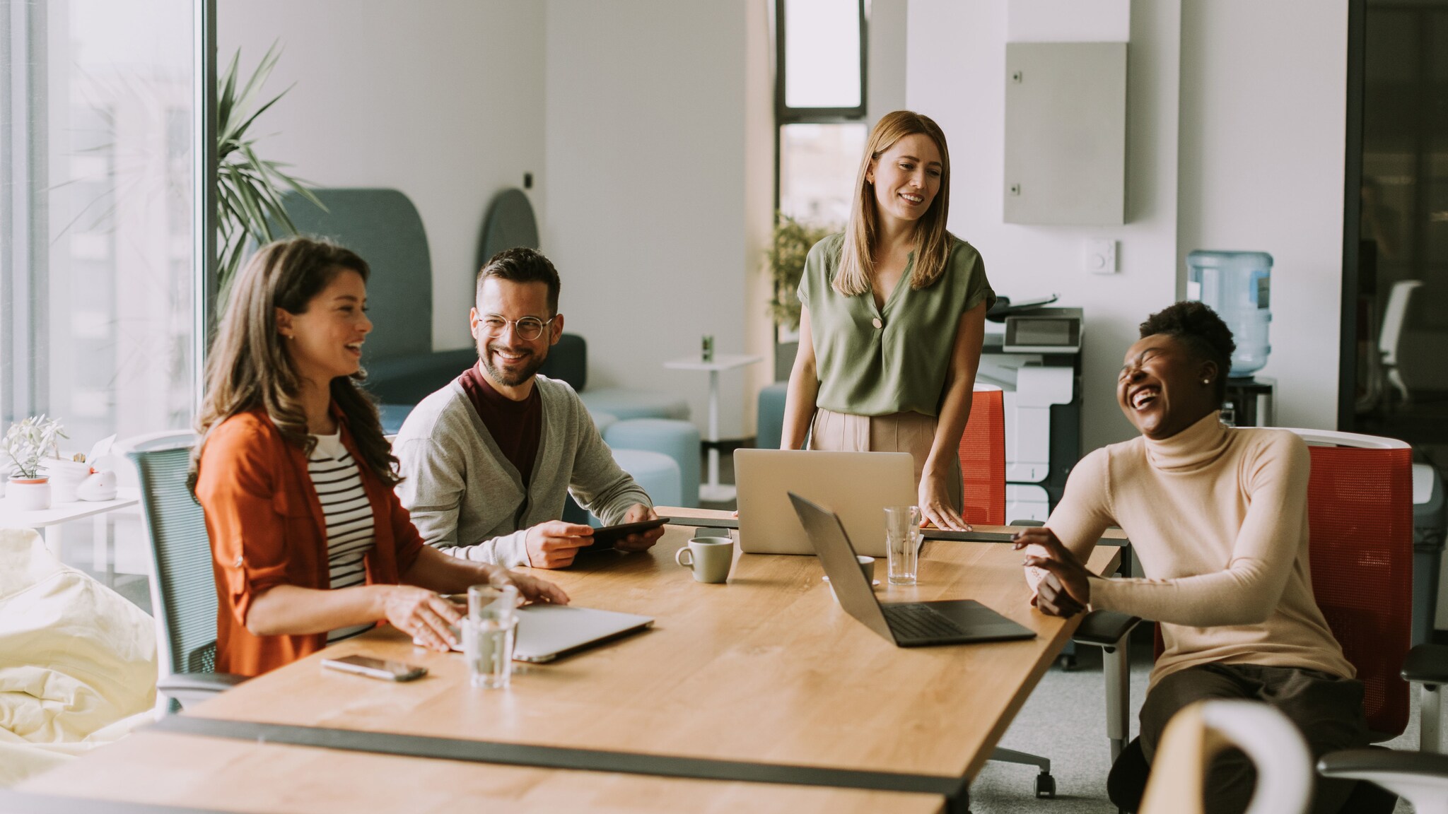 Vier lächelnde Personen sitzen, eine steht, an einem Konferenztisch mit Laptops.