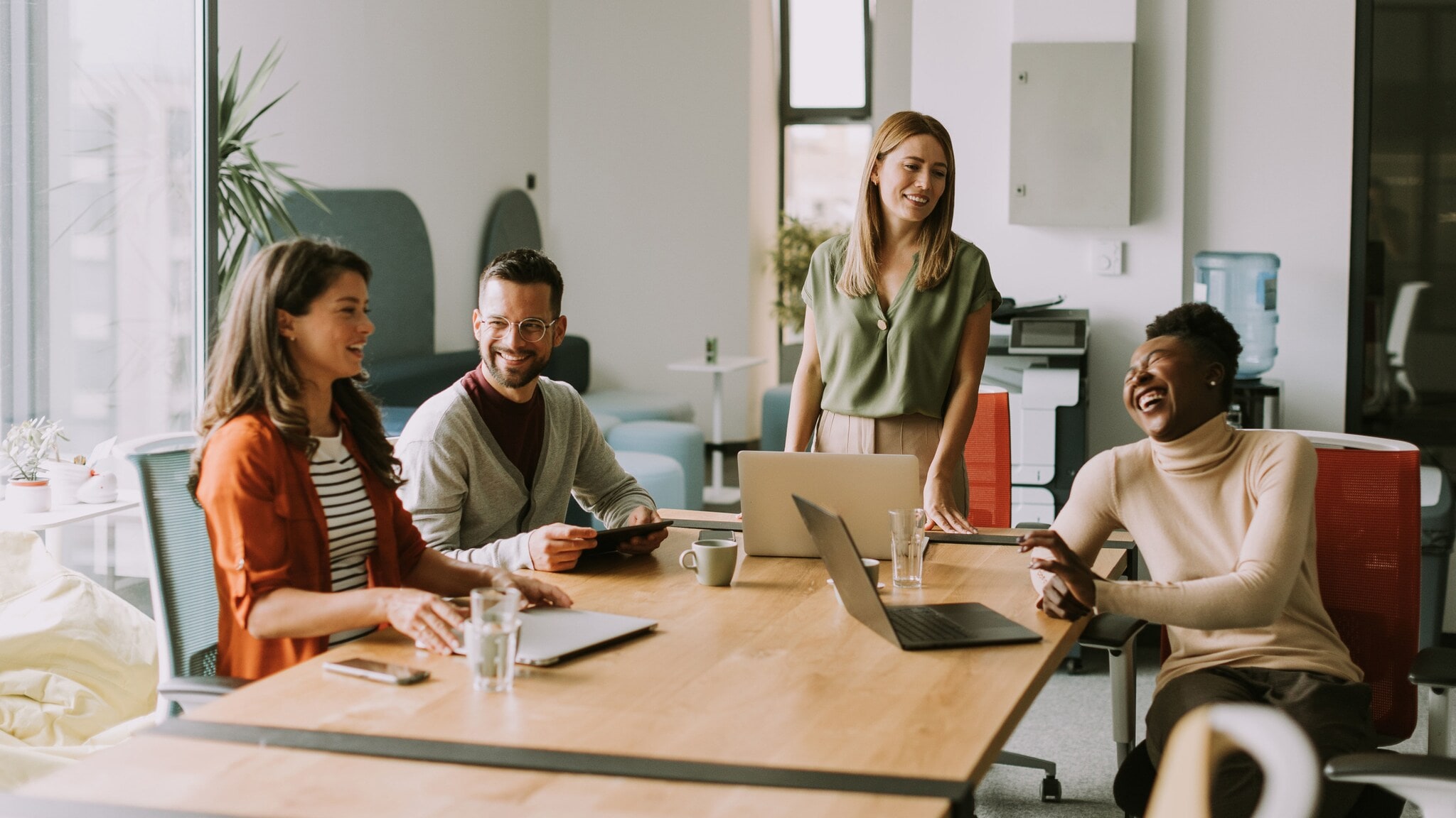Vier lächelnde Personen sitzen, eine steht, an einem Konferenztisch mit Laptops. Vier lächelnde Personen sitzen, eine steht, an einem Konferenztisch mit Laptops.