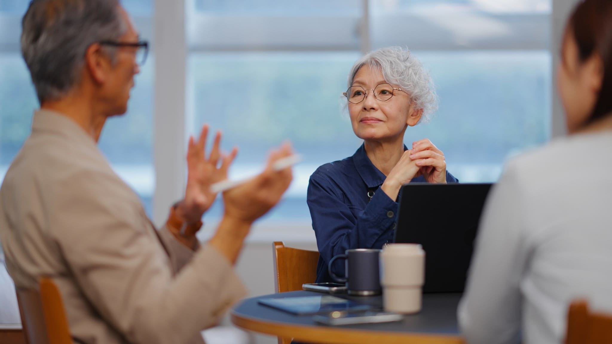 Eine ältere asiatische Frau mit grauen Locken sitzt mit Laptop an einem Tisch mit zwei weiteren Personen, von der eine mit den Händen gestikuliert. Eine ältere asiatische Frau mit grauen Locken sitzt mit Laptop an einem Tisch mit zwei weiteren Personen, von der eine mit den Händen gestikuliert.