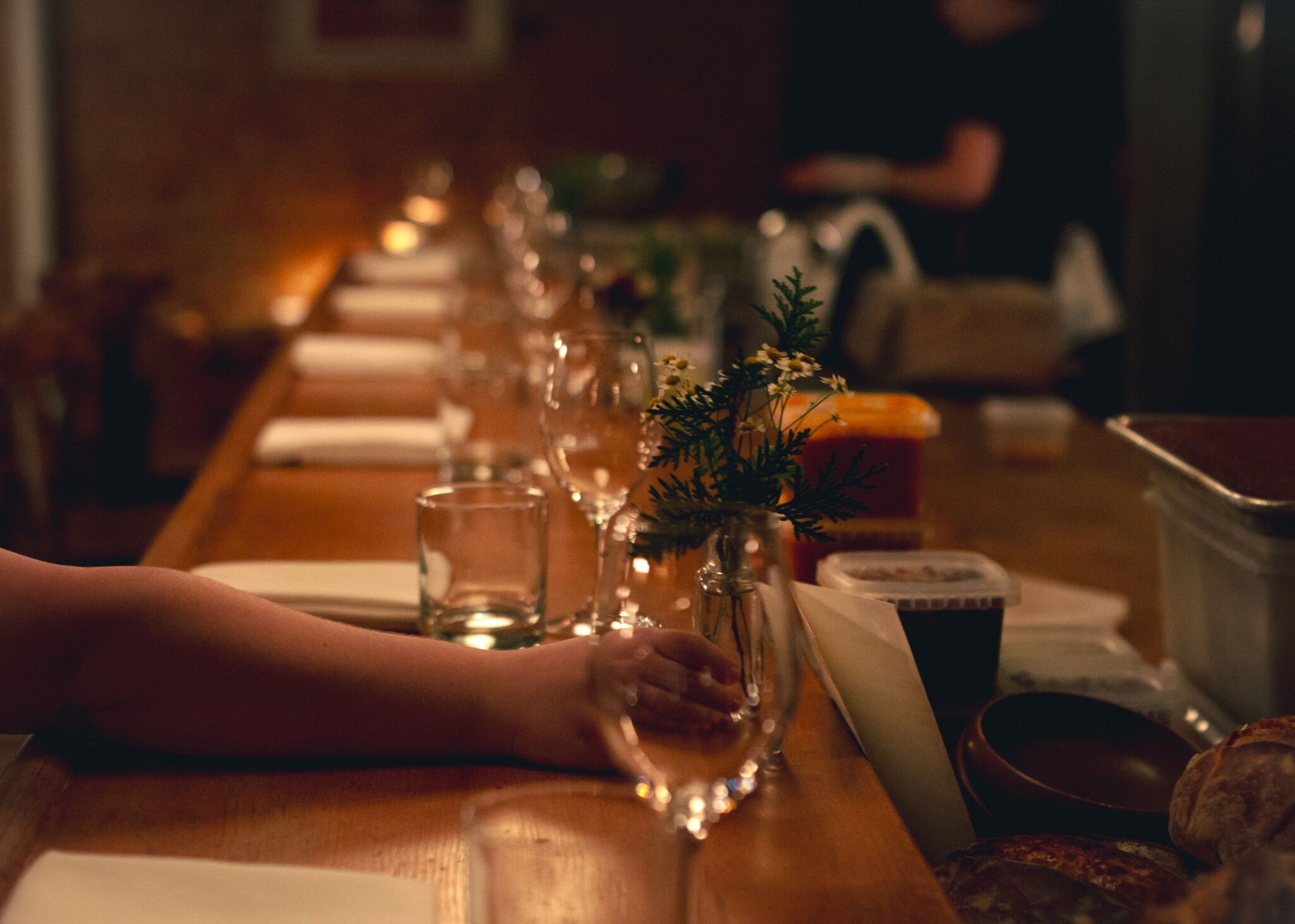 Gedeckter Holztresen bei schummrigen Licht, auf den eine Frauenhand eine kleine Blumenvase mit Kamille stellt.