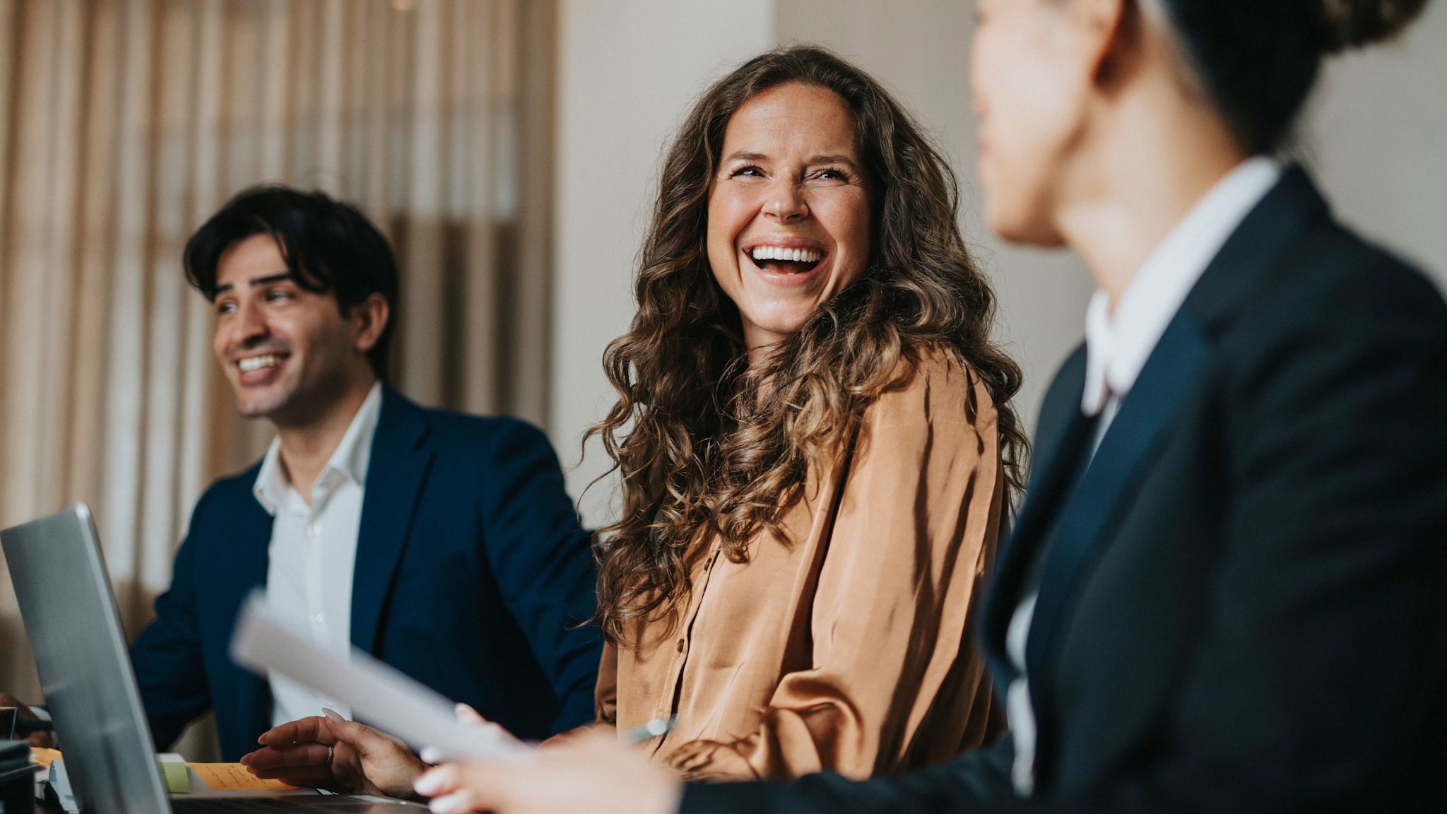 Eine lachende Frau sitzt vor einem Laptop an einem Tisch zwischen zwei weiteren Personen in Business-Kleidung während eines Meetings. Eine lachende Frau sitzt vor einem Laptop an einem Tisch zwischen zwei weiteren Personen in Business-Kleidung während eines Meetings.