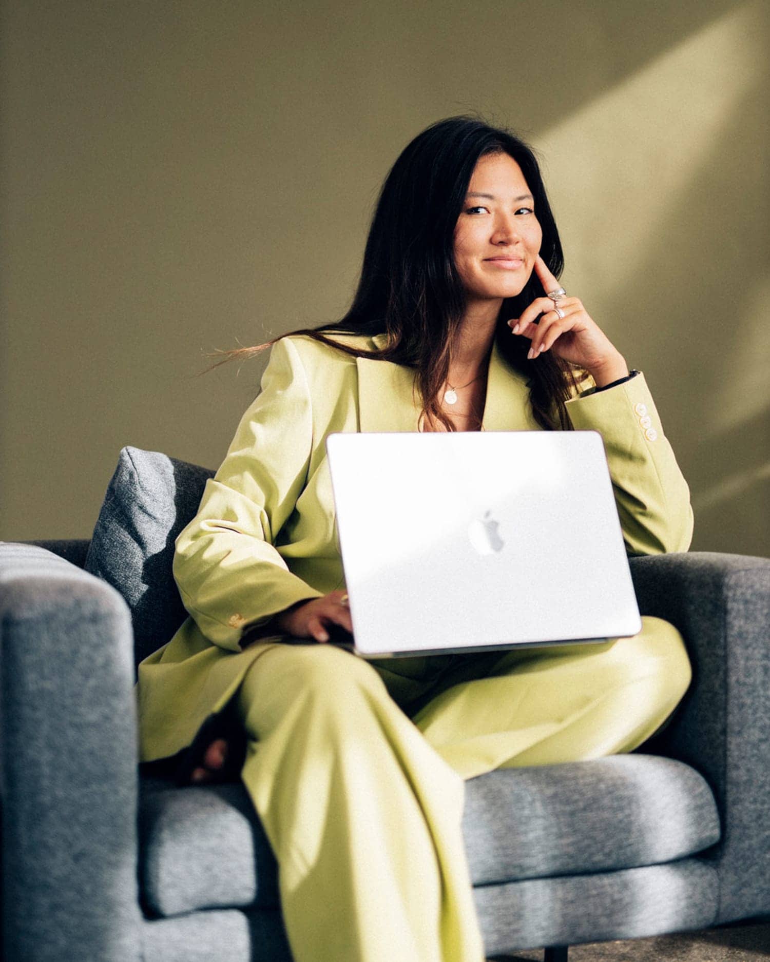 ine Frau sitzt in einem hellgelben Anzug mit aufgestütztem Arm und aufgeklapptem Laptop auf dem Schoß in einem Sessel und lächelt dezent in die Kamera. ine Frau sitzt in einem hellgelben Anzug mit aufgestütztem Arm und aufgeklapptem Laptop auf dem Schoß in einem Sessel und lächelt dezent in die Kamera.