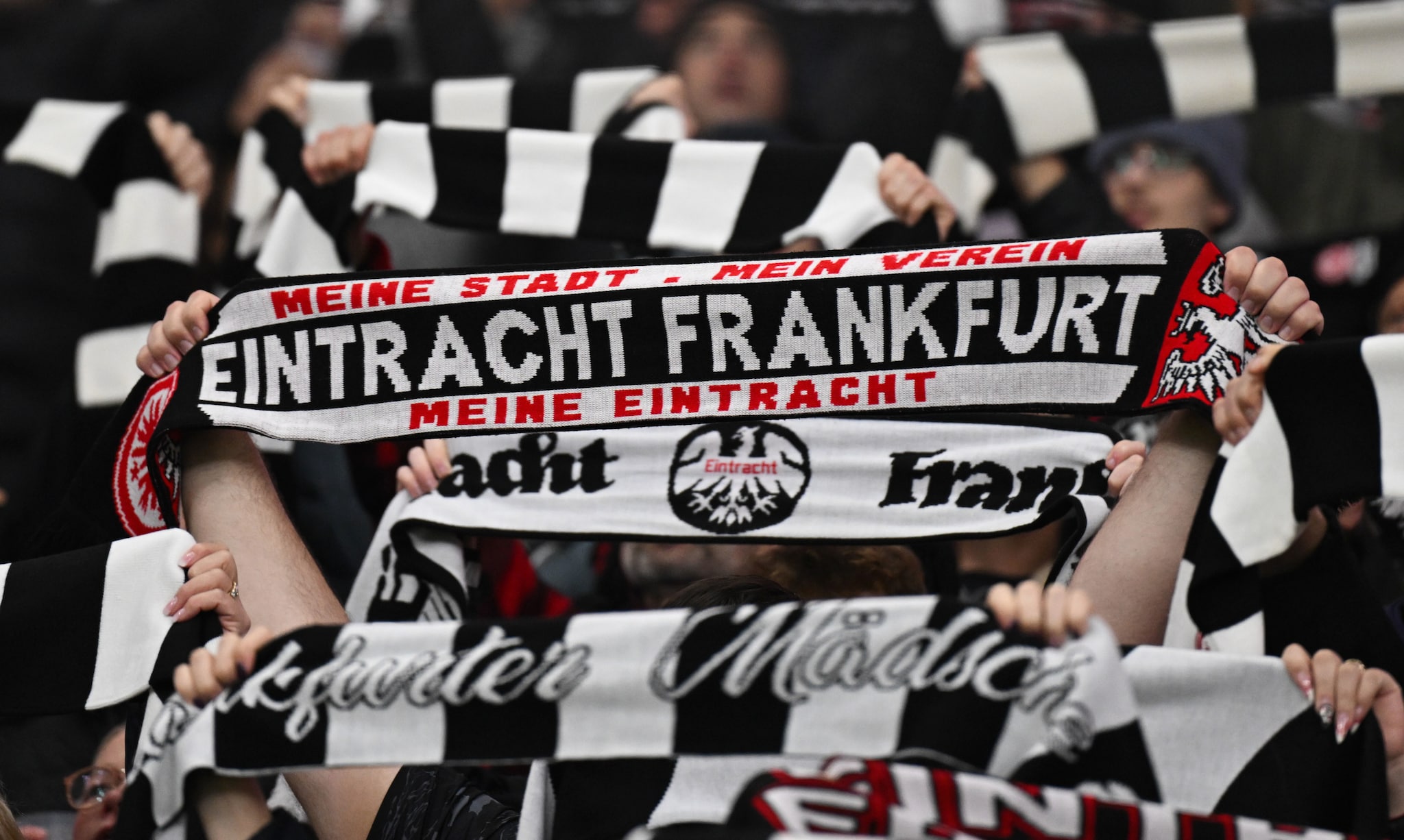Personen halten schwarz-weiße Schals mit Eintracht-Frankfurt-Schriftzug über ihre Köpfe auf einer Tribüne.