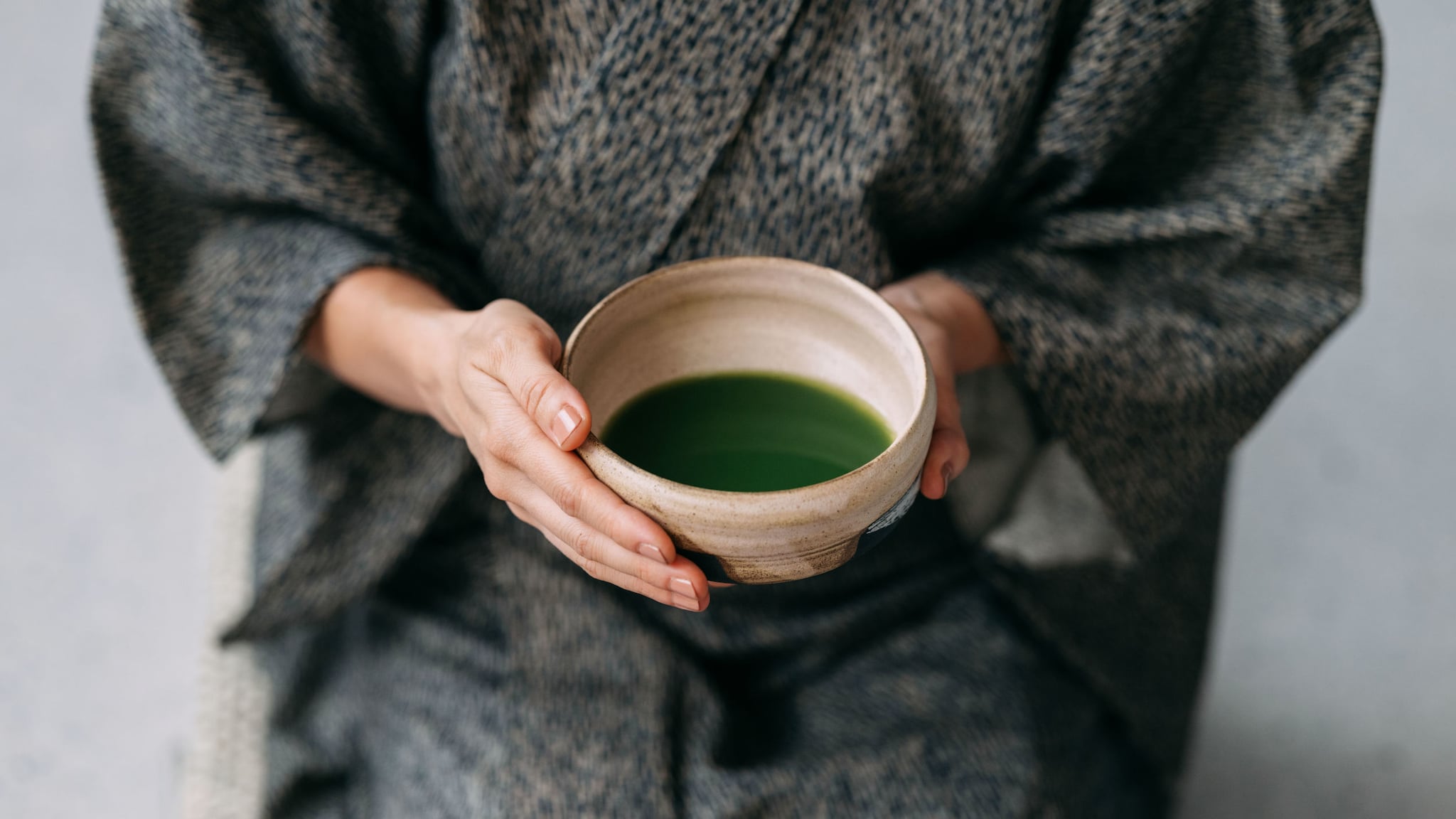 Nahaufnahme einer Keramikschale mit grünem Getränk in den Händen einer sitzenden Person im grauen Kimono.