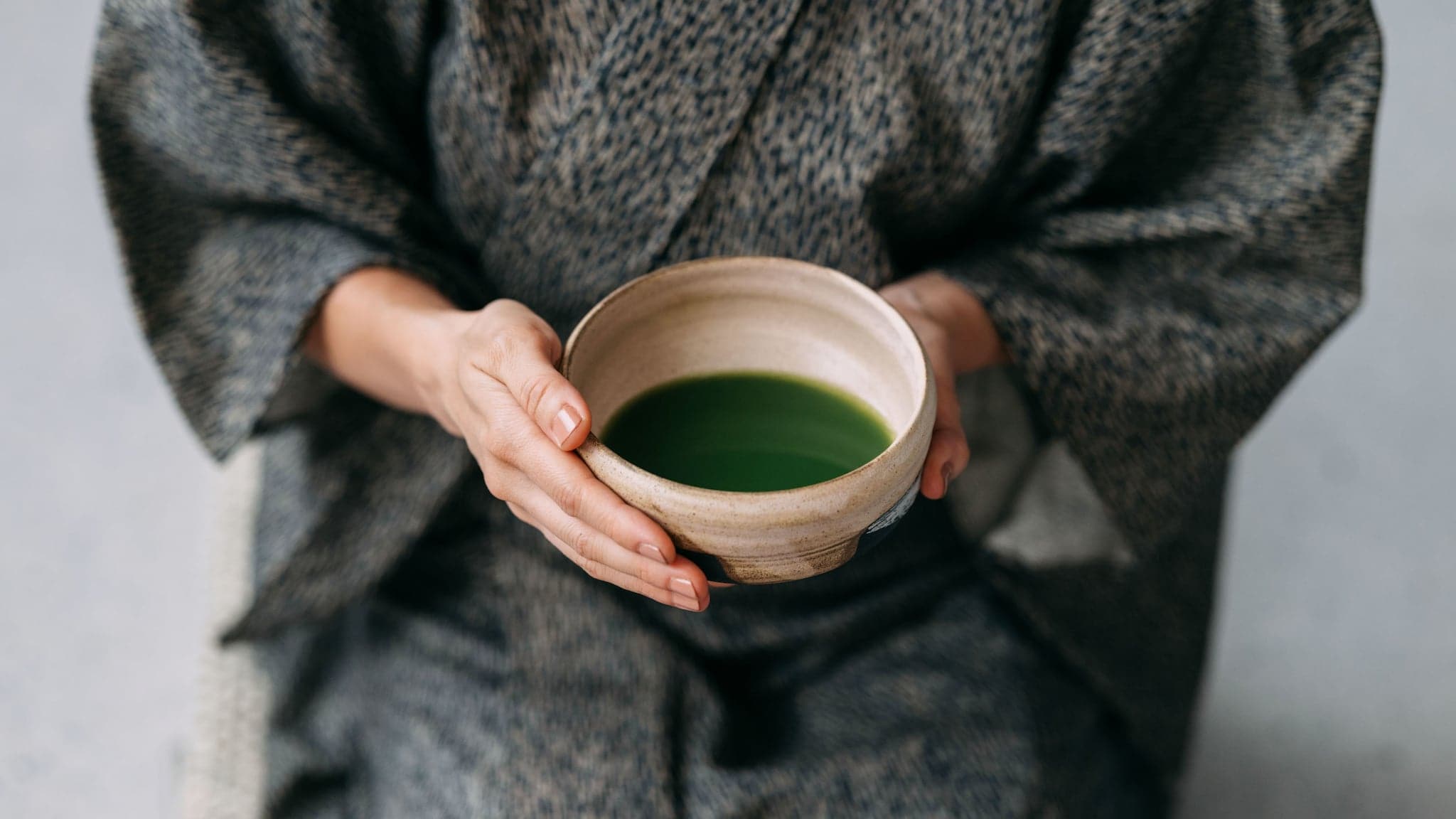 Nahaufnahme einer Keramikschale mit grünem Getränk in den Händen einer sitzenden Person im grauen Kimono. Nahaufnahme einer Keramikschale mit grünem Getränk in den Händen einer sitzenden Person im grauen Kimono.