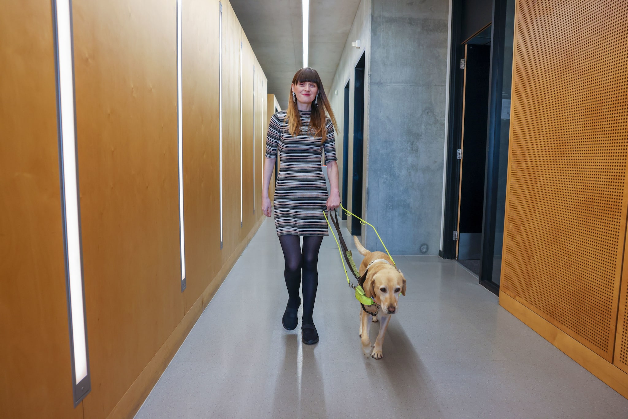 Eine junge Frau im Kleid geht mit einem Blindenhund einen Flur entlang.