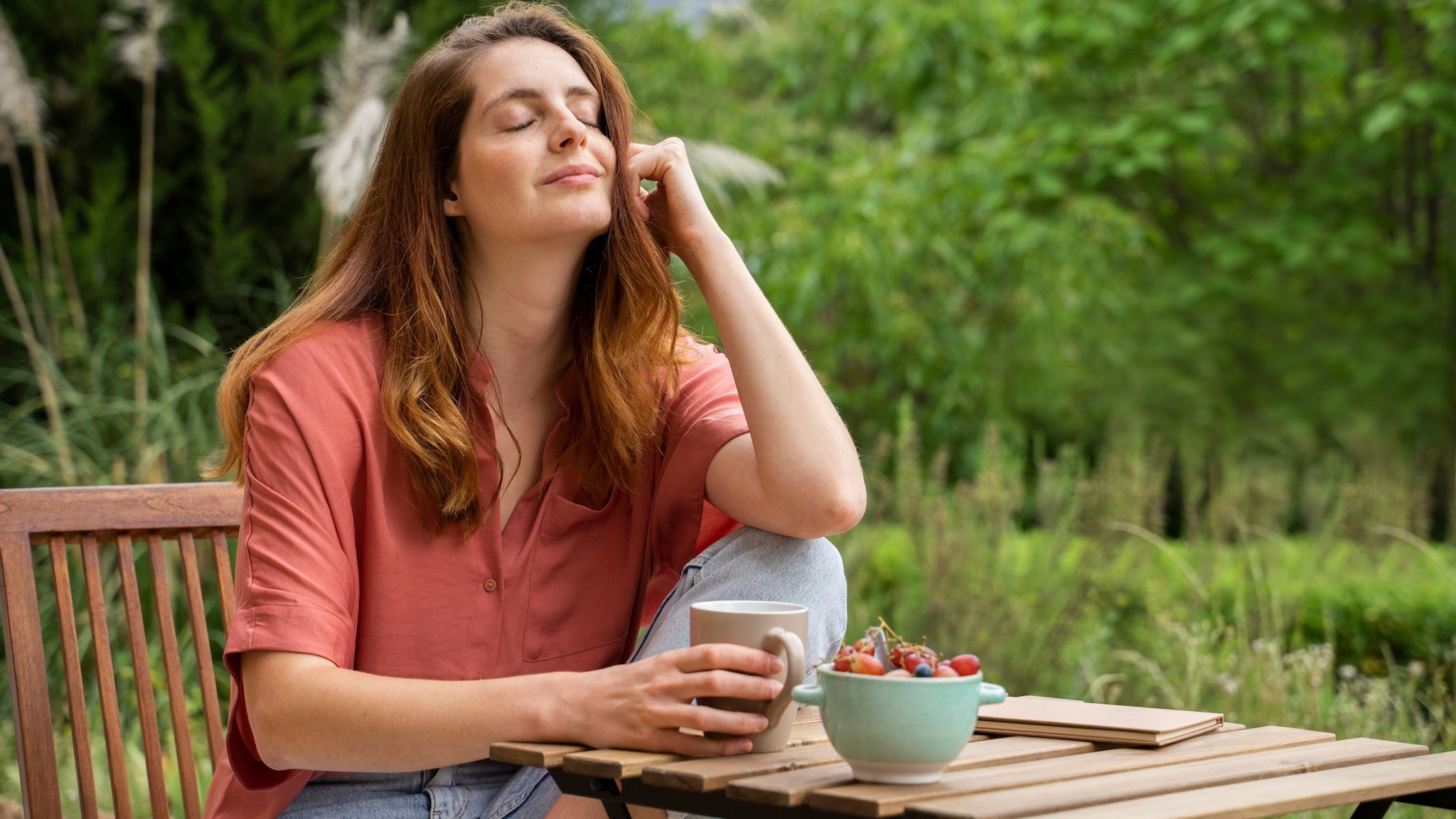 Eine entspannt lächelnde Frau mit geschlossenen Augen sitzt mit Kaffeebecher und angewinkeltem Bein an einem Holztisch in einem Garten. Eine entspannt lächelnde Frau mit geschlossenen Augen sitzt mit Kaffeebecher und angewinkeltem Bein an einem Holztisch in einem Garten.