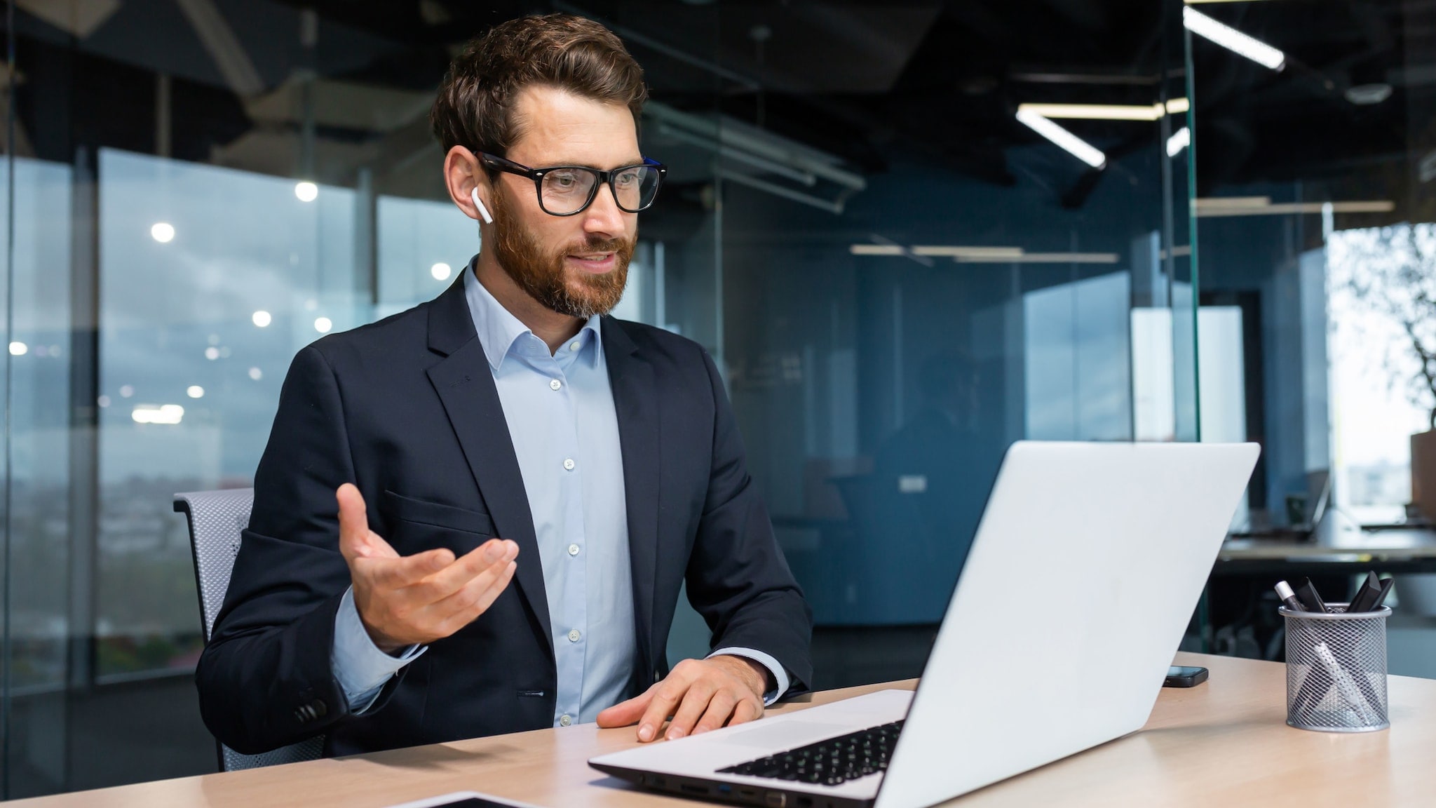 Ein Mann im Anzug und Kopfhörern in den Ohren sitzt an einem Schreibtisch vor einem aufgeklappten Laptop und gestikuliert mit der Hand in einem Raum mit Glaswand.