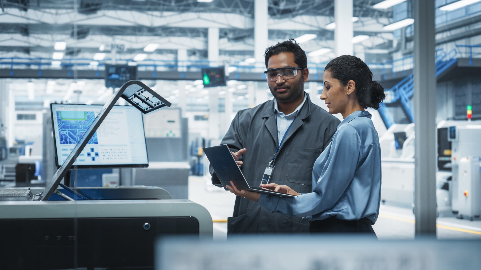 Ein Mann und eine Frau stehen an einer digitalen Schaltzentrale einer Elektrofabrik und schauen gemeinsam auf ein Laptop.