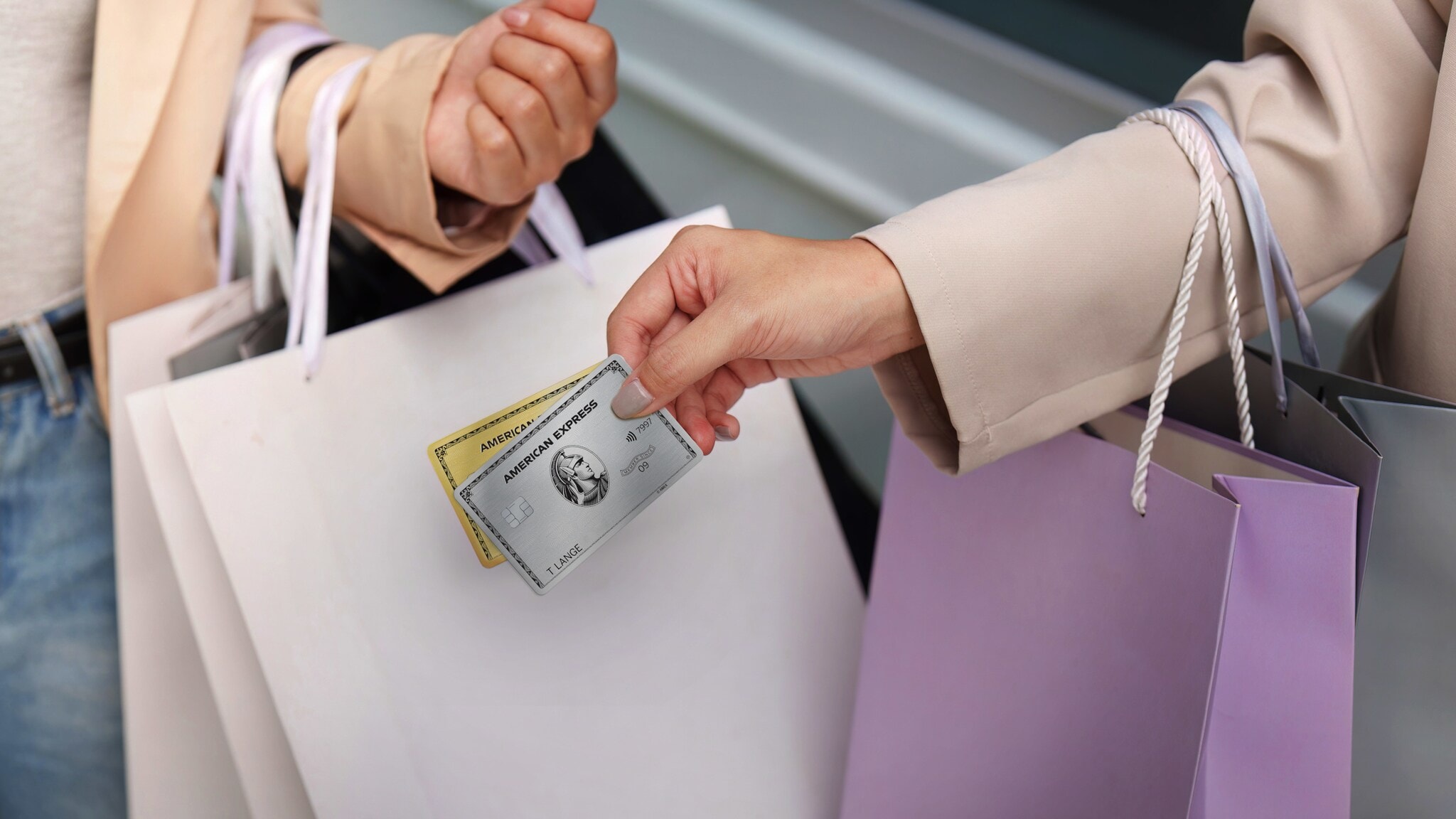 Nahaufnahme von zwei Armen mit Einkaufstüten, eine Hand hält eine goldene und eine silberne Kreditkarte von American Express. Nahaufnahme von zwei Armen mit Einkaufstüten, eine Hand hält eine goldene und eine silberne Kreditkarte von American Express.