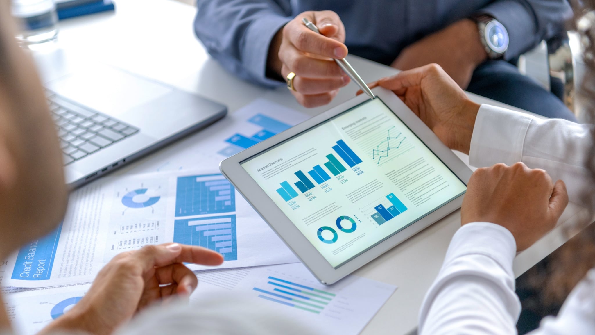 Drei Personen sitzen an einem Tisch, auf dem Zettel mit Diagrammen liegen. Eine Person hält ein Tablet, das Diagramme zeigt. Drei Personen sitzen an einem Tisch, auf dem Zettel mit Diagrammen liegen. Eine Person hält ein Tablet, das Diagramme zeigt.