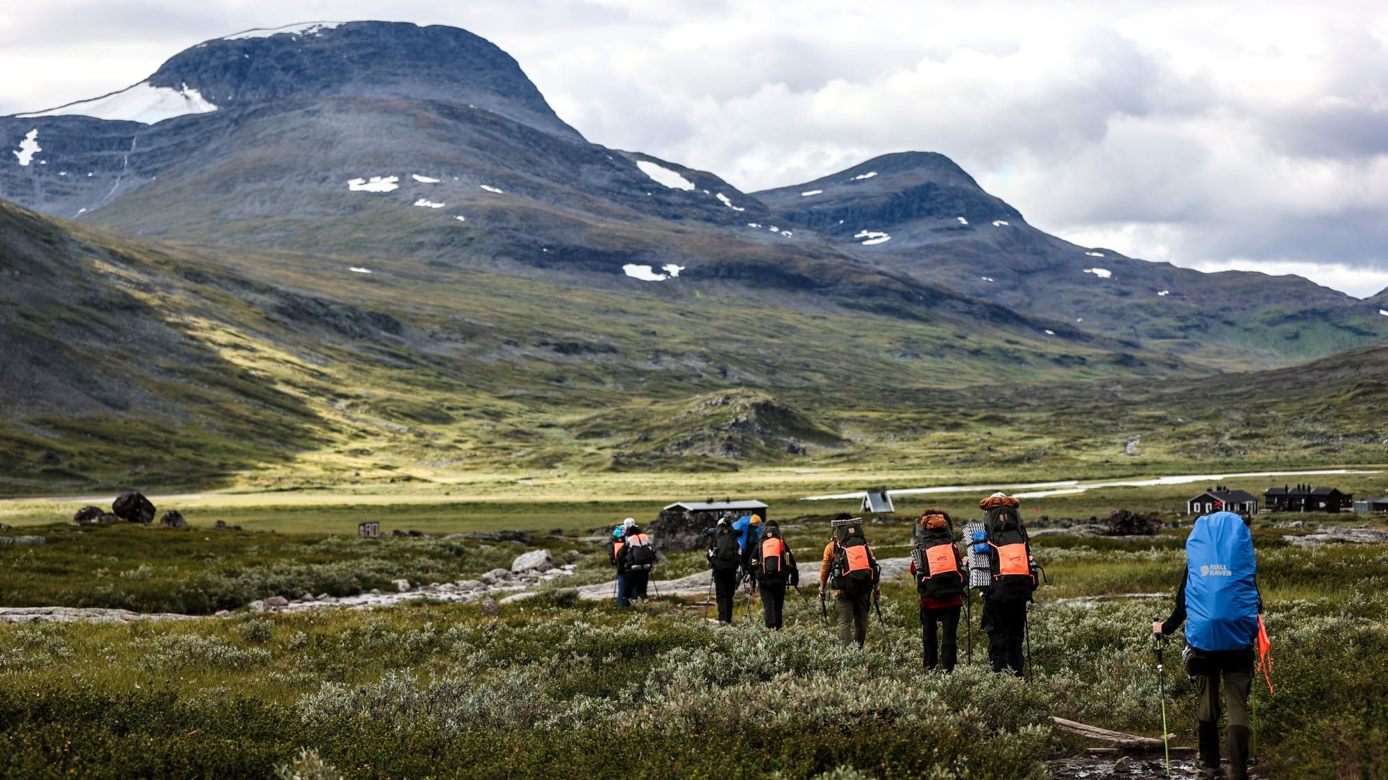 Gruppe von sieben Wanderern mit Rucksäcken auf einem Wanderweg in einer bergigen Landschaft. Im Hintergrund sind hohe Berge und bewölkter Himmel sichtbar. Gruppe von sieben Wanderern mit Rucksäcken auf einem Wanderweg in einer bergigen Landschaft. Im Hintergrund sind hohe Berge und bewölkter Himmel sichtbar.