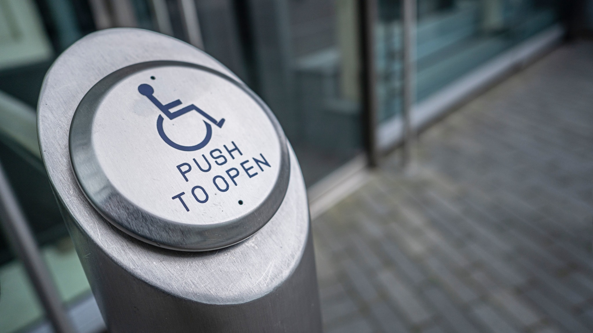 Nahaufnahme eines Pfeilers mit Knopf mit der Aufschrift push to open und einem Rollstuhlsymbol vor einem Gebäudeeingang. Nahaufnahme eines Pfeilers mit Knopf mit der Aufschrift push to open und einem Rollstuhlsymbol vor einem Gebäudeeingang.