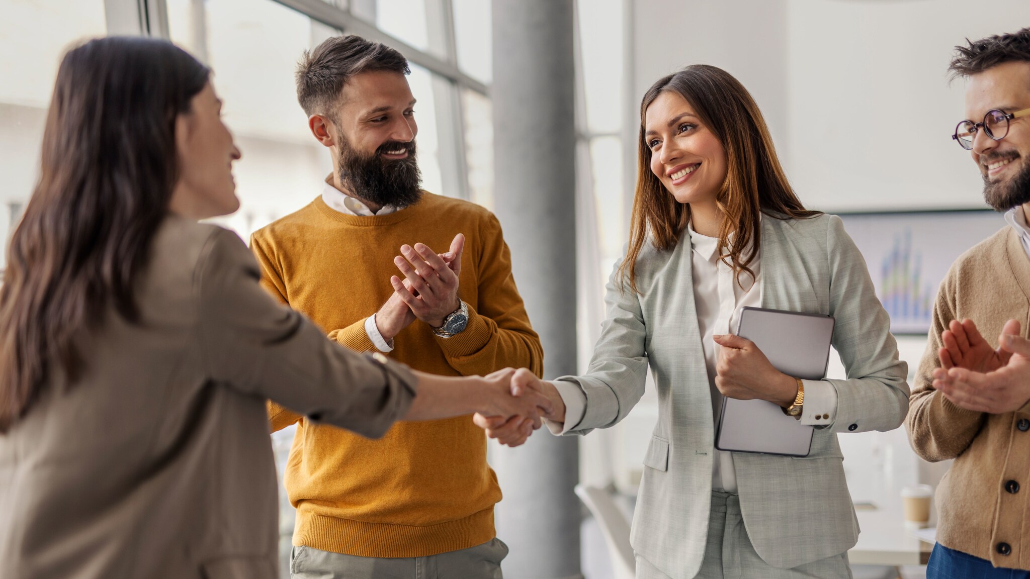 Vier Personen stehen beieinander, zwei Frauen geben sich lächelnd die Hand, während zwei Männer in die Hände klatschen.