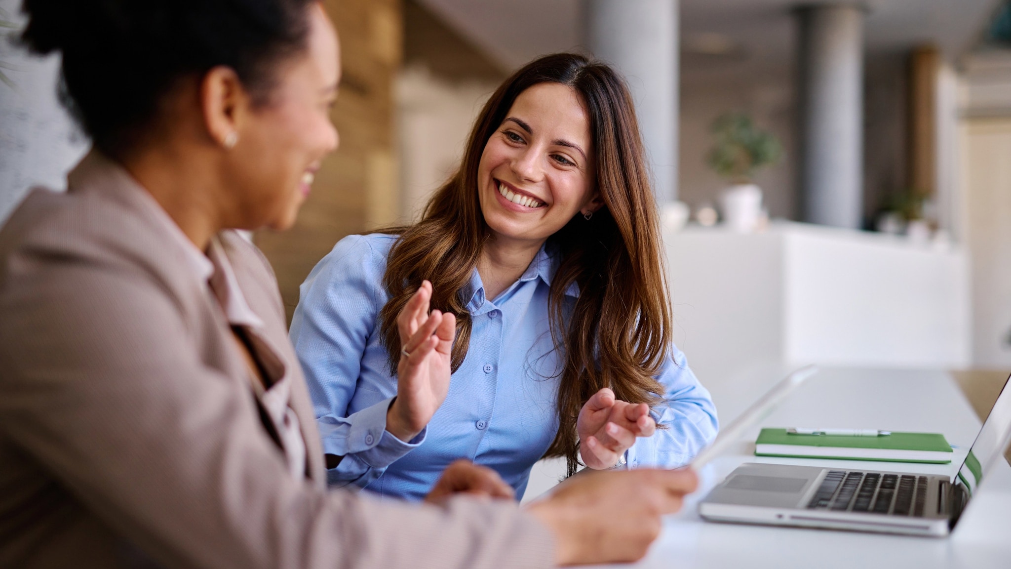 Zwei Frauen sitzen nebeneinander an einem Tisch, auf dem ein aufgeklappter Laptop steht und ein Notizbuch liegt, und lächeln sich an. Zwei Frauen sitzen nebeneinander an einem Tisch, auf dem ein aufgeklappter Laptop steht und ein Notizbuch liegt, und lächeln sich an.